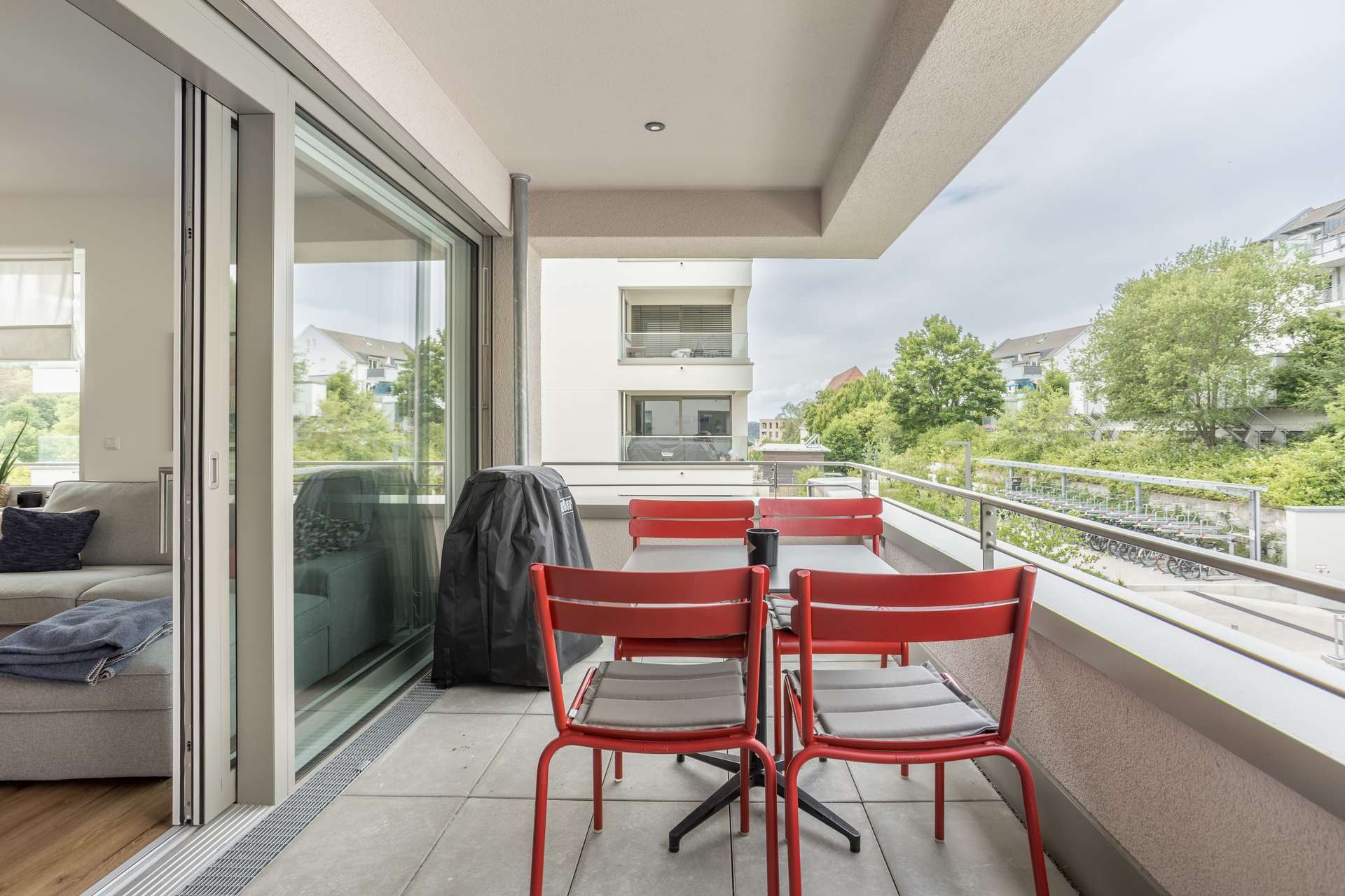 Balkon Ein moderner Balkon mit einem roten Tisch und vier roten Stühlen, einem Grill, einem Glasgeländer und einem Blick auf die umliegenden Wohnhäuser und Bäume. Glasschiebetüren führen in ein helles Wohnzimmer.