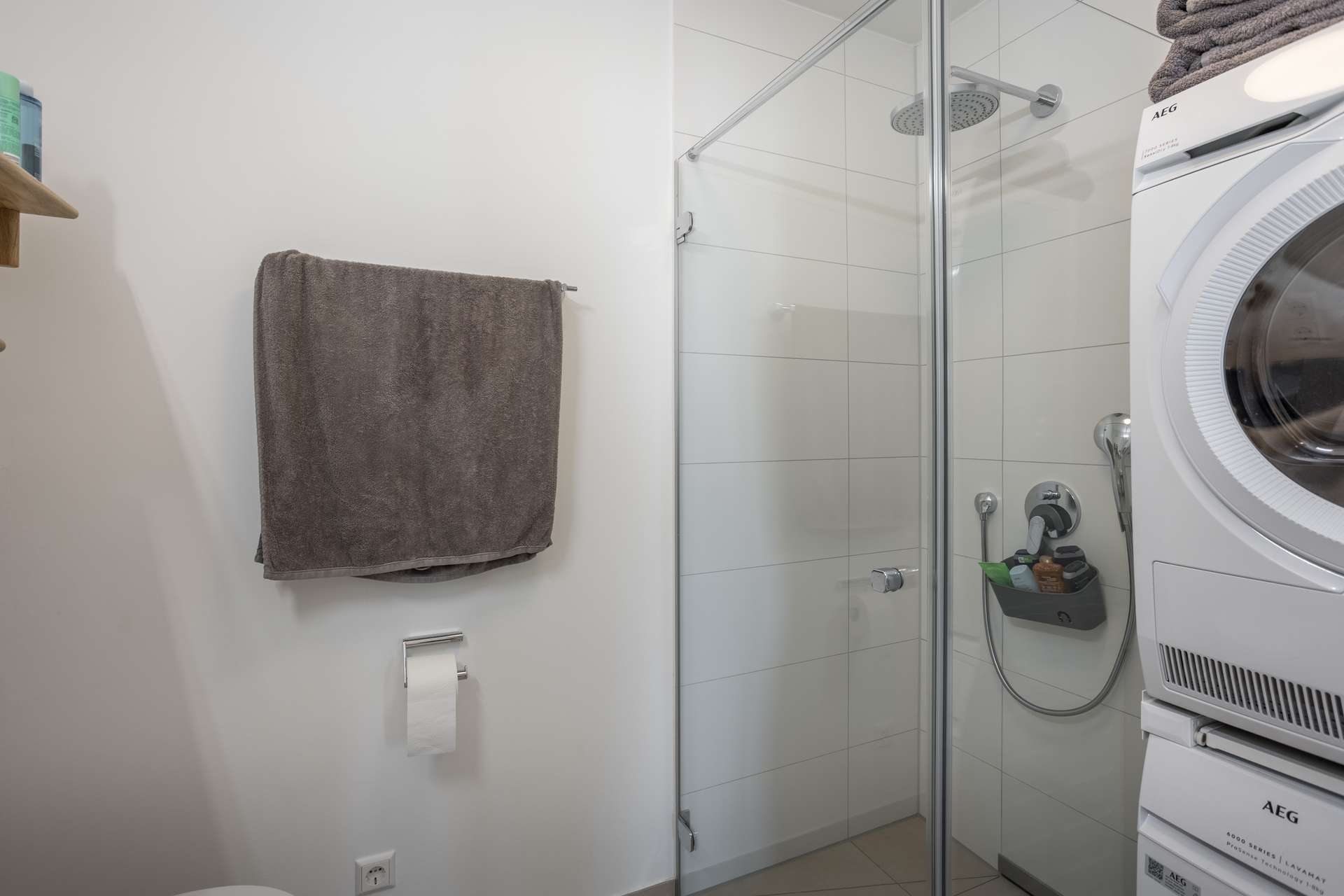 Badezimmer Modernes Badezimmer mit weiß gefliester Dusche, Glastür, grauen Handtüchern an der Wand, Toilettenpapierhalter und einer gestapelten Wasch- und Trockeneinheit auf der rechten Seite. Die Regale mit Toilettenartikeln sind teilweise sichtbar.