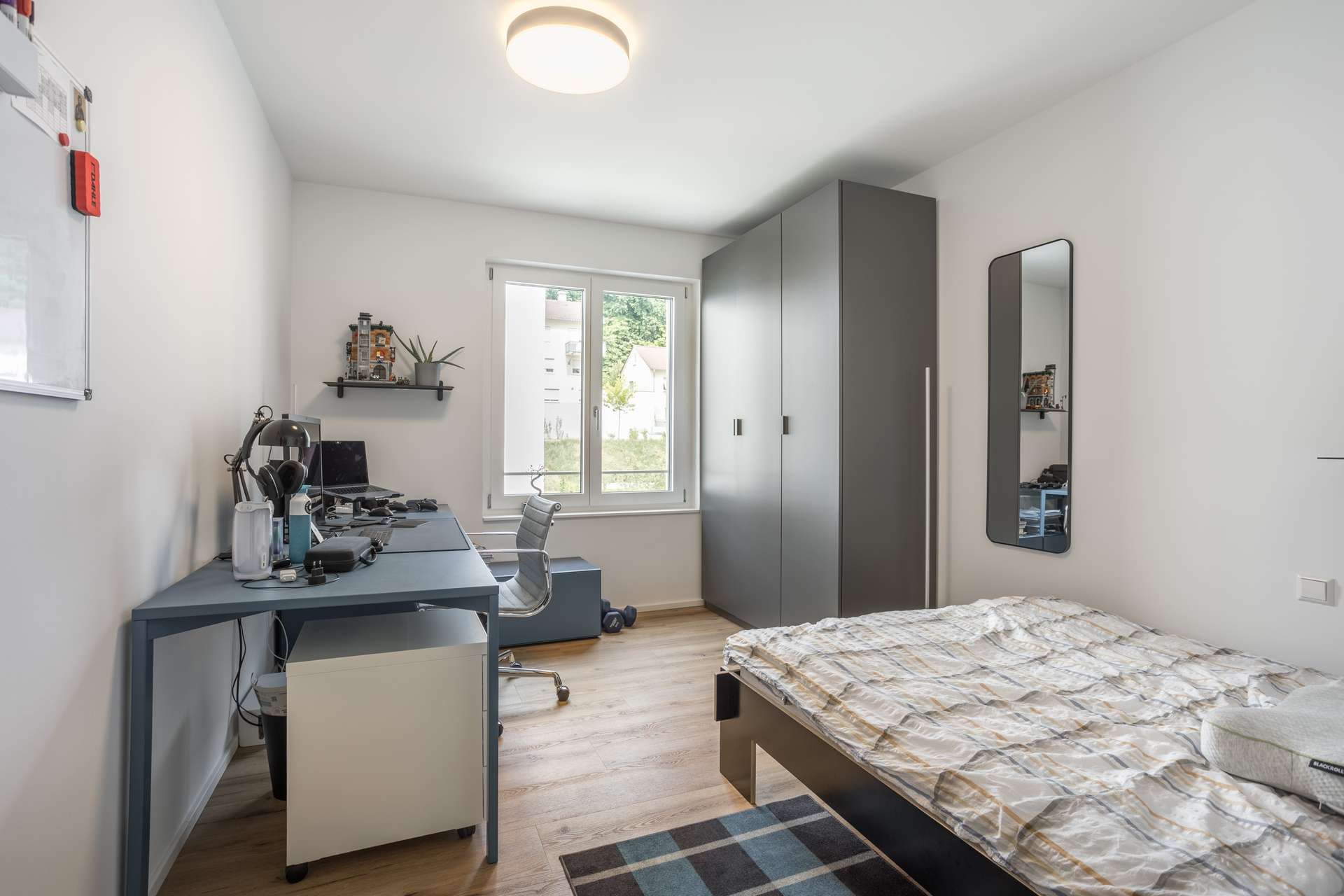 Schlafzimmer 2 Ein modernes, aufgeräumtes Schlafzimmer mit einem Einzelbett, einem grauen Kleiderschrank, einem Wandspiegel und einem Schreibtisch mit einem Computer. Ein Fenster lässt natürliches Licht herein, und der Boden ist aus hellem Holz mit einem kleinen Teppichboden.