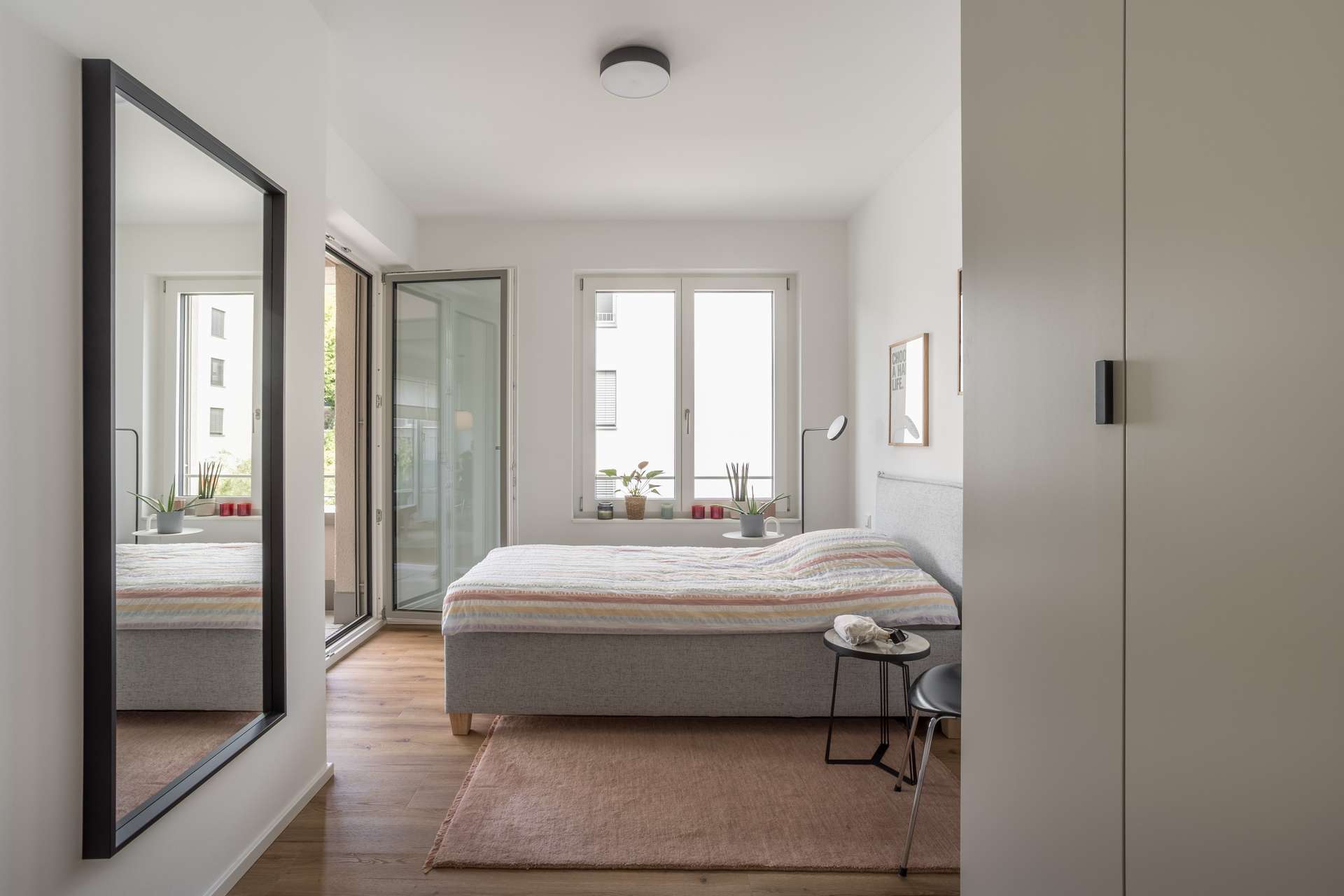 Schlafzimmer 1 Helles, modernes Schlafzimmer mit großem Bett, Nachttisch, Pflanzen auf der Fensterbank, Wandspiegel, Glastür zum Balkon und Holzfußboden mit rosa Teppich. Minimalistisches Dekor und weiße Wände sorgen für ein luftiges Gefühl.
