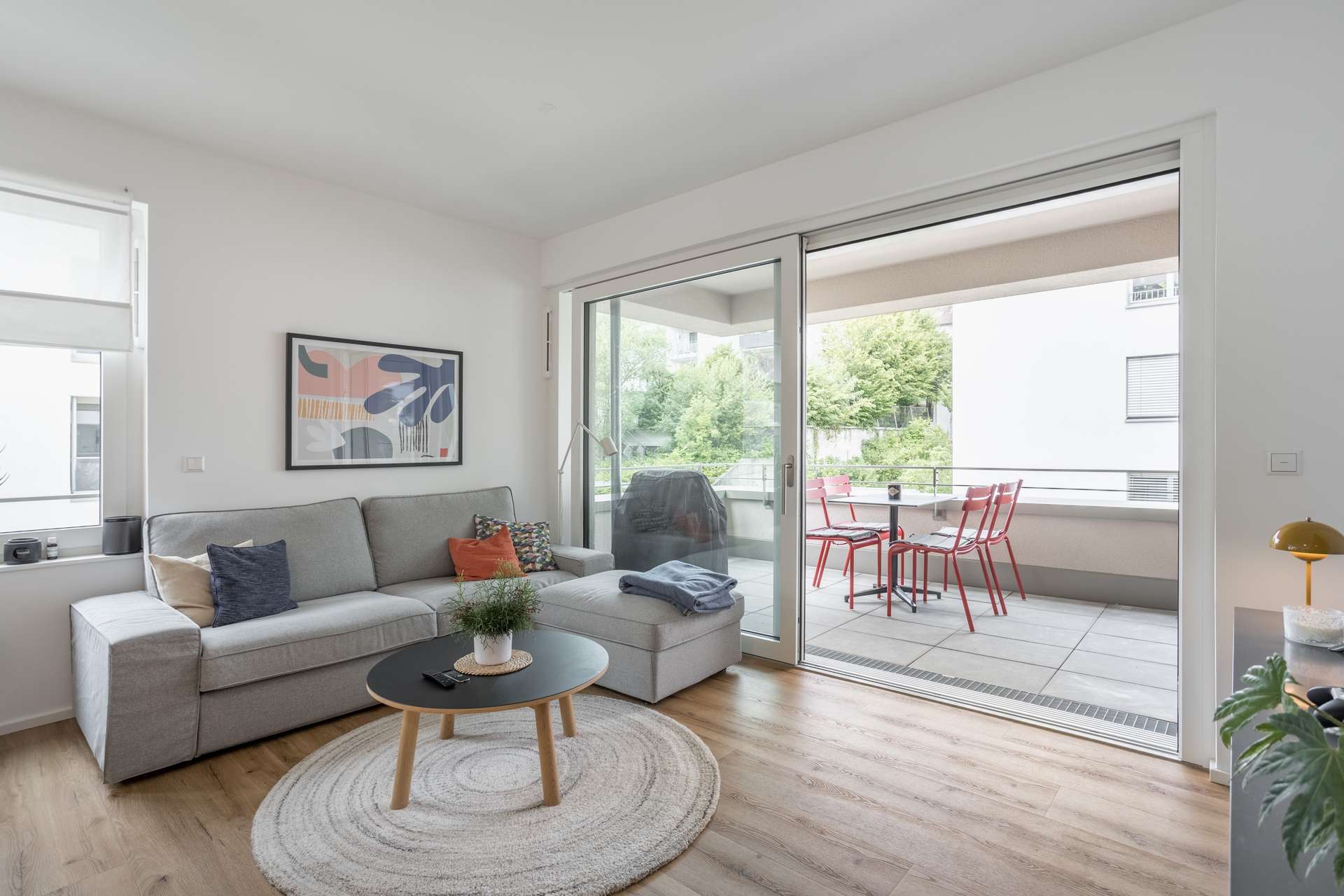 Wohn- und Essbereich Ein modernes Wohnzimmer mit einem grauen Sektionssofa, einem Couchtisch und abstrakter Kunst. Große Glasschiebetüren führen auf einen Balkon mit einem Tisch und roten Stühlen, von dem aus man ins Grüne blickt. Der Raum ist hell und luftig und hat helle Holzböden.