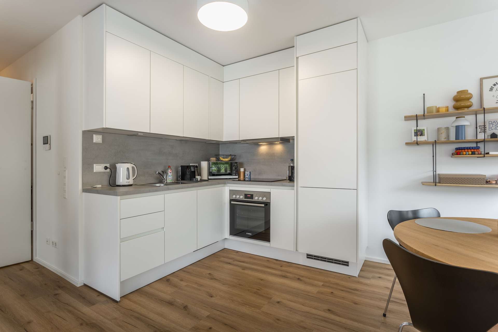 Küche Moderne Küche mit weißen Schränken, grauer Aufkantung, eingebautem Ofen und verschiedenen Kleingeräten. Es gibt einen runden Esstisch mit schwarzen Stühlen und Regale mit Dekorationsartikeln an der Wand auf der rechten Seite. Holzfußboden im ganzen Haus.