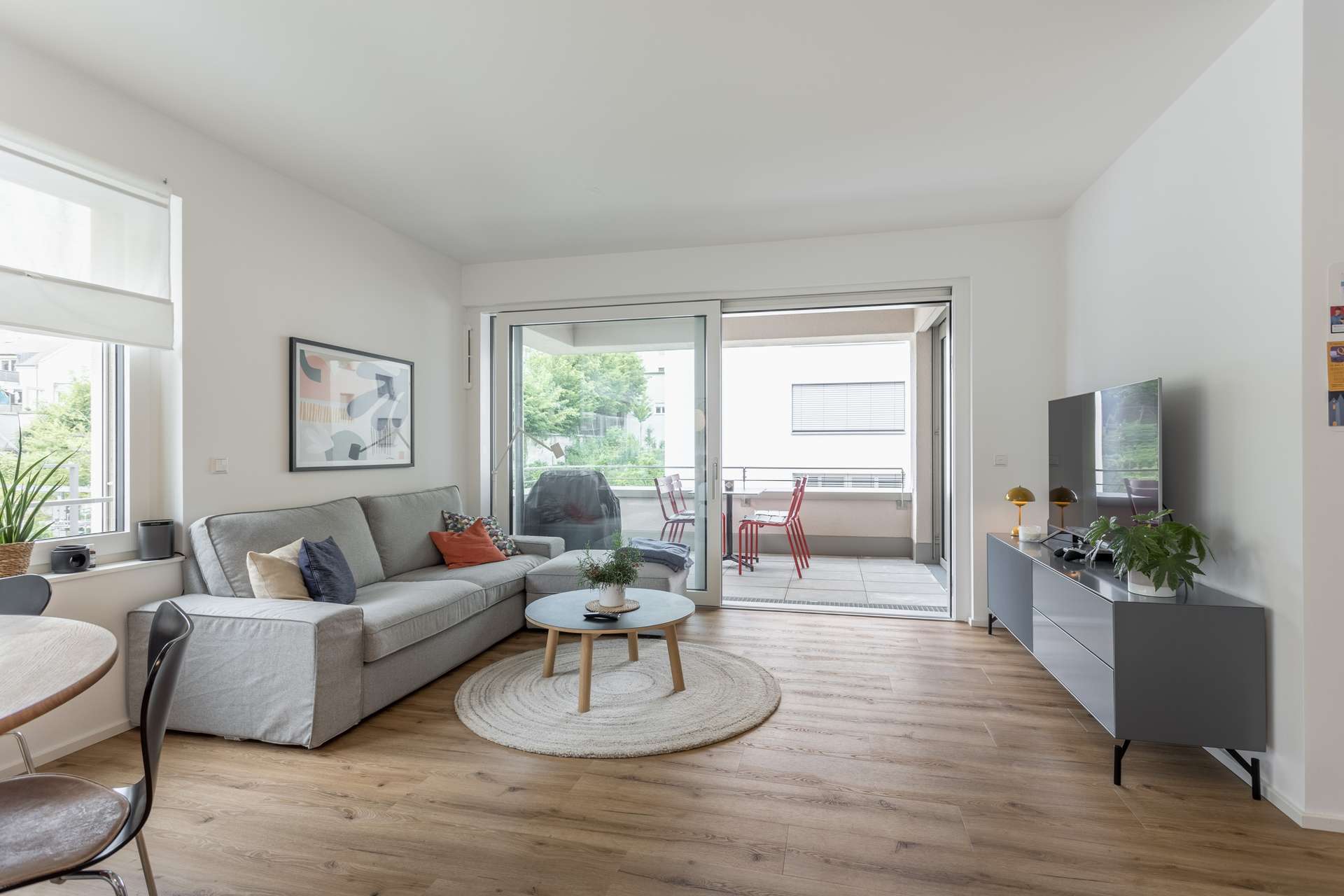 Wohn- und Essbereich Modernes Wohnzimmer mit einem grauen Sofa, einem runden Couchtisch, einem Fernseher auf einer Konsole und großen Glasschiebetüren, die auf einen Balkon mit einem Tisch und roten Stühlen führen. Der Raum hat einen Holzboden und ist hell mit natürlichem Licht.