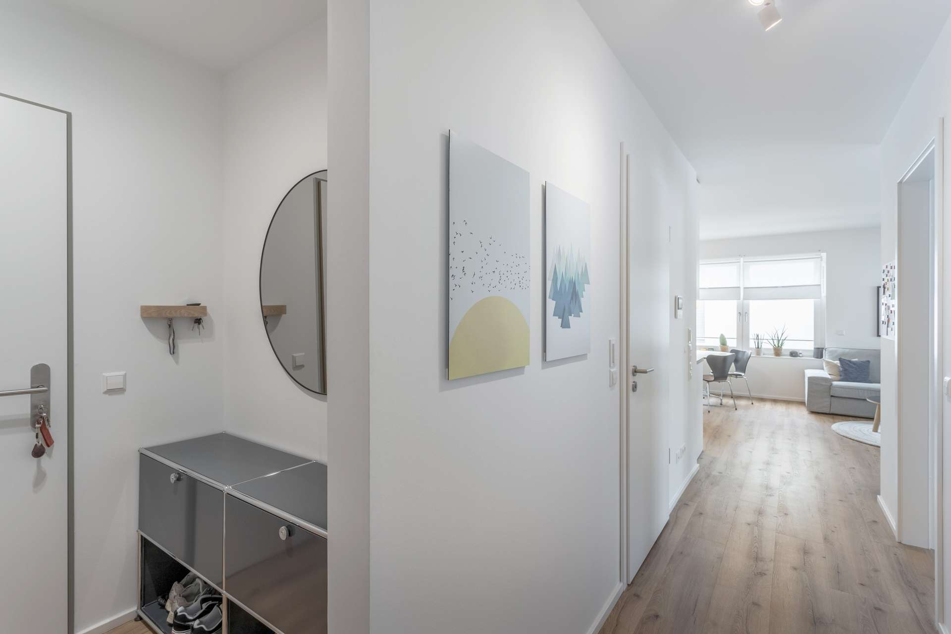 Eingangsbereich / Flur Ein heller, moderner Flur mit hellen Wänden und Holzböden. Auf der linken Seite befindet sich ein runder Spiegel über den Ablageschränken. An der rechten Wand hängen zwei minimalistische Kunstdrucke, die in ein sonnendurchflutetes Wohnzimmer führen.