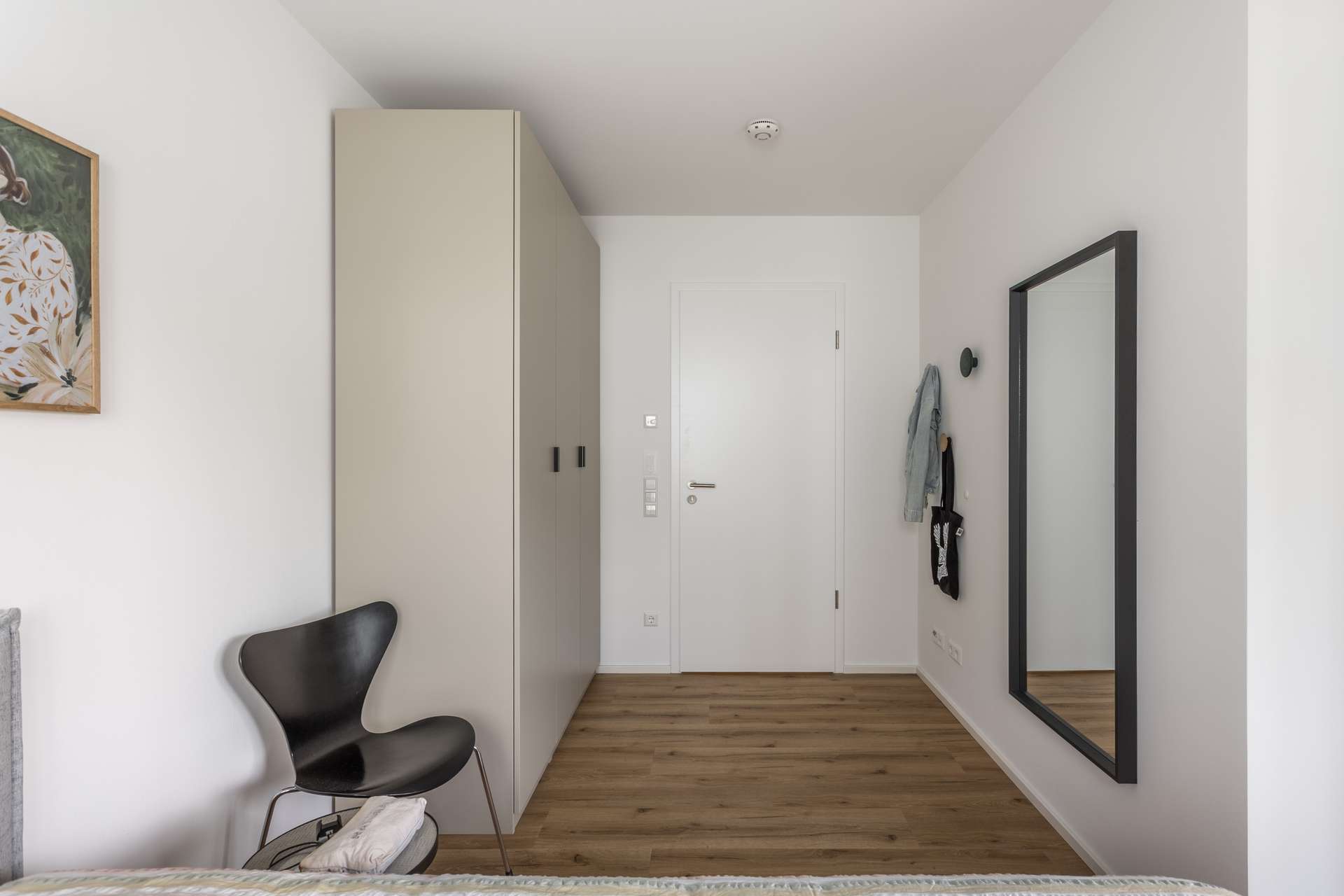 Schlafzimmer 1 Minimalistisches Schlafzimmer mit weißen Wänden, einem großen Kleiderschrank, einem schwarzen Stuhl, einem Wandspiegel, einem kleinen Gemälde und ein paar an Haken hängenden Gegenständen. Der Boden ist aus hellem Holz und die Tür ist geschlossen.