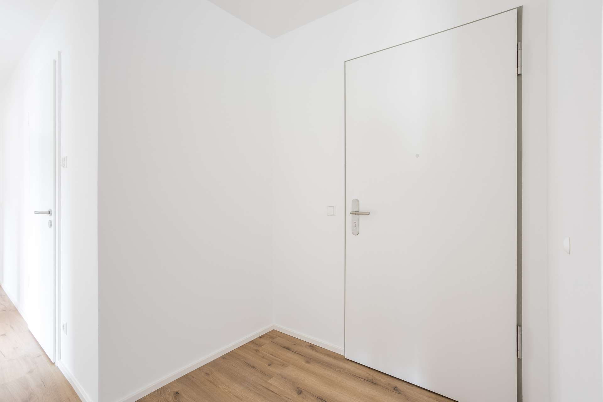 Ansicht_Eingang Ein minimalistisch gestalteter Flur mit weißen Wänden, einem hellen Holzboden und einer großen weißen Tür mit silbernem Türgriff. Weiter hinten auf der linken Seite des Flurs ist eine weitere weiße Tür zu sehen.