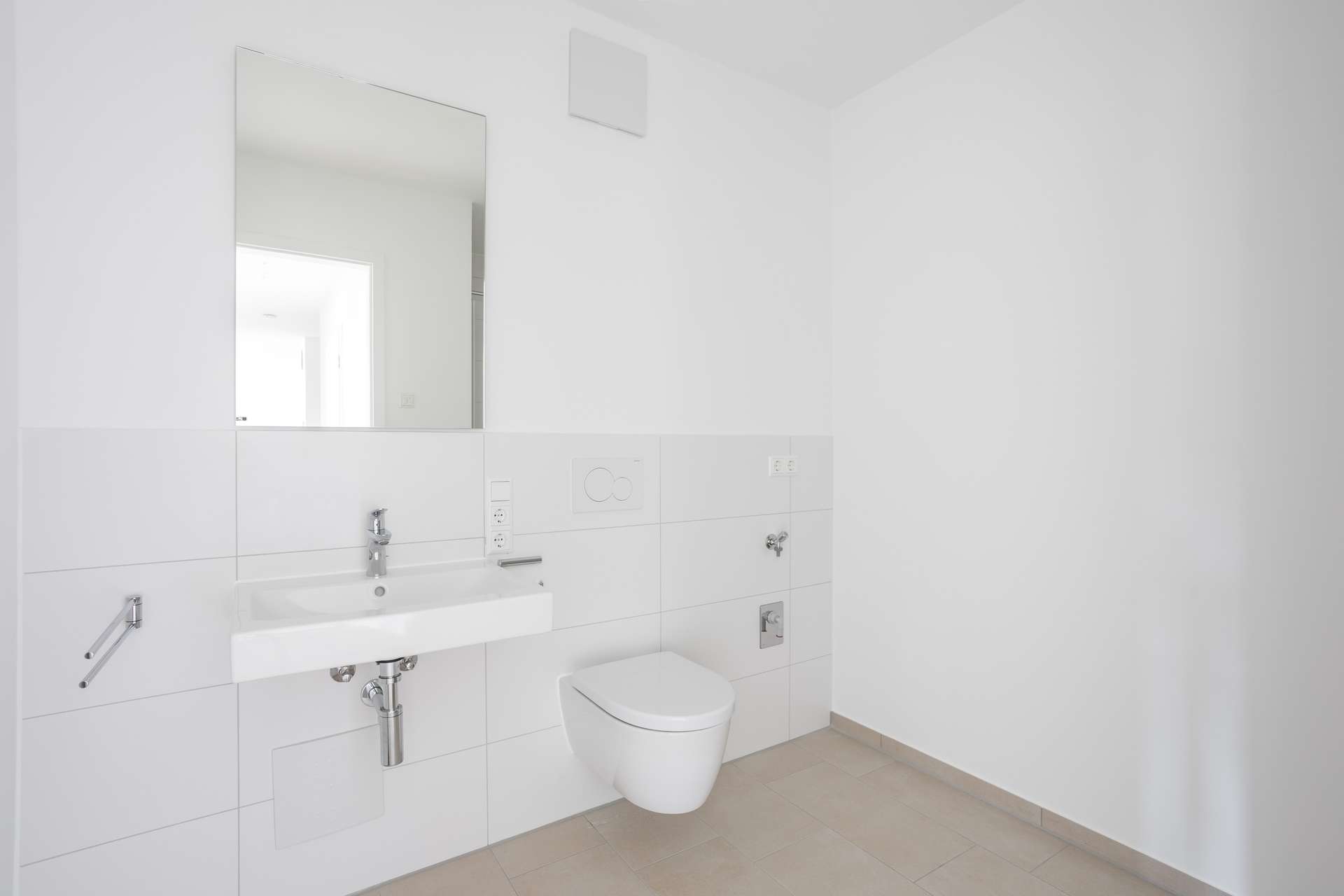 Ansicht_Bad Ein minimalistisches Badezimmer mit weißen Wänden, einem rechteckigen Spiegel über einem weißen Waschbecken, einem wandhängenden WC und hellbeigen Bodenfliesen. Der Raum wirkt hell und aufgeräumt und ist mit modernen Armaturen ausgestattet.