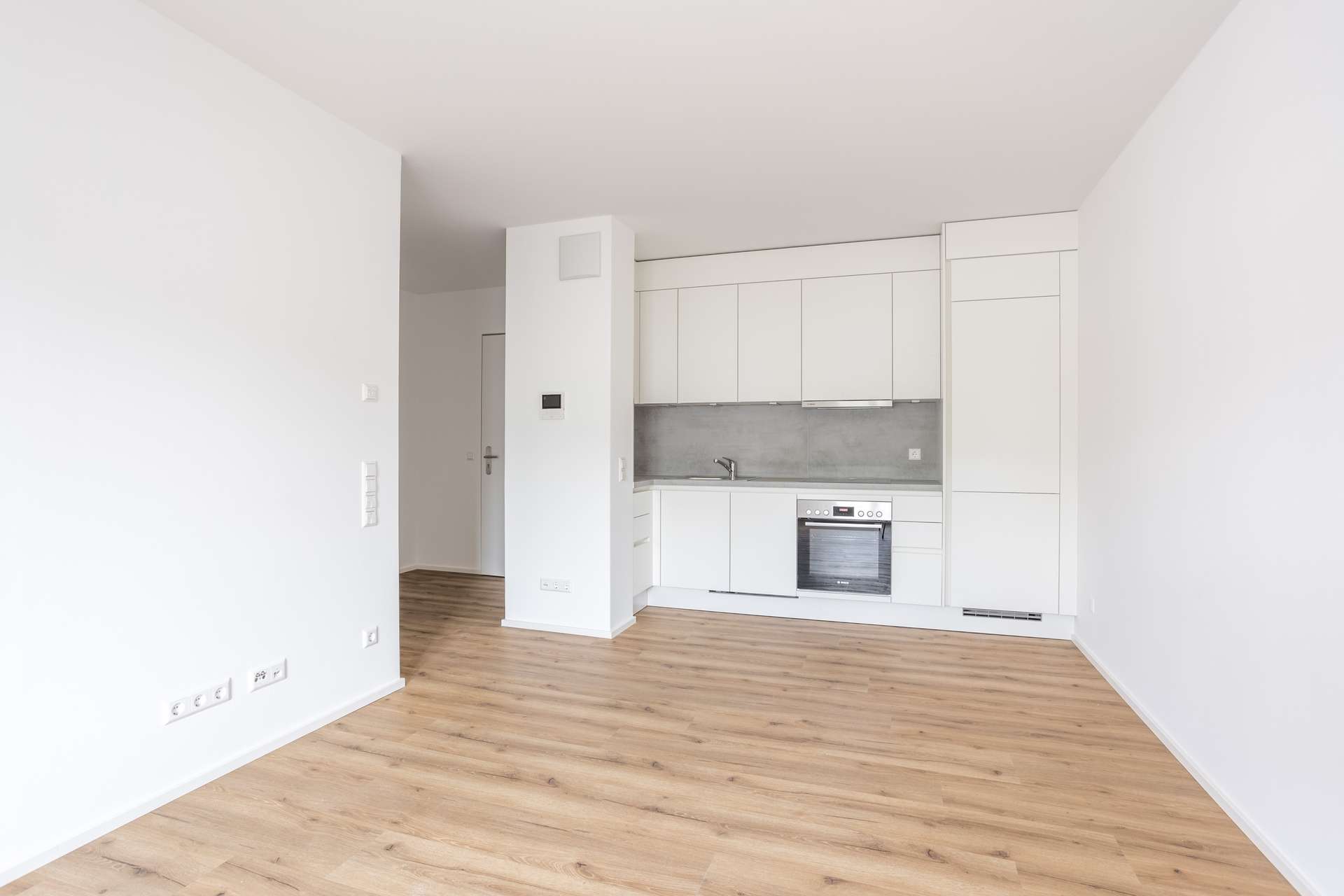 Ansicht_Küche Helle, moderne, unmöblierte Wohnung mit weißen Wänden, hellem Holzboden und einer kompakten Küche mit weißen Schränken, einer Spüle, einem Kochfeld und einem Einbaubackofen. Der Raum ist sauber, minimalistisch und aufgeräumt.