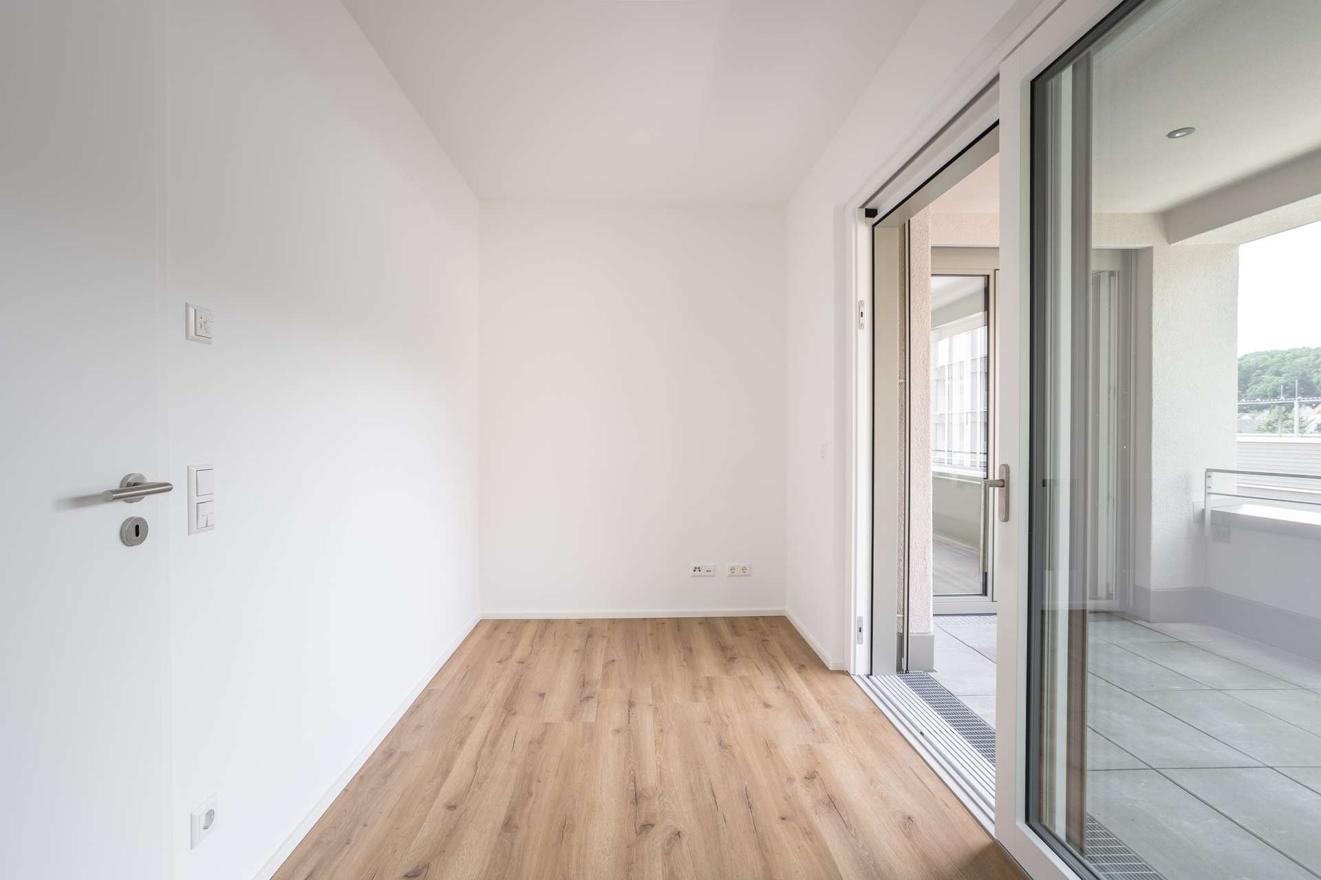 Ansicht_Gast_Büro Ein kleiner, leerer Raum mit hellem Holzboden, weißen Wänden und einer Glasschiebetür, die auf einen überdachten Balkon mit Blick auf Bäume und Gebäude im Freien führt.