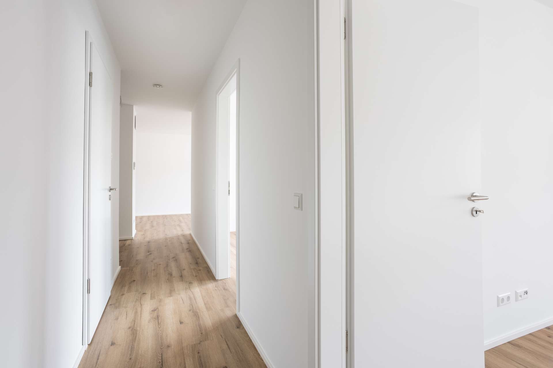 Ansicht_Flur Heller Flur in einem modernen Haus mit weißen Wänden, offenen weißen Türen und hellem Holzboden. Mehrere Türöffnungen führen zu verschiedenen Räumen, und natürliches Licht durchflutet den Raum und schafft eine klare und großzügige Atmosphäre.