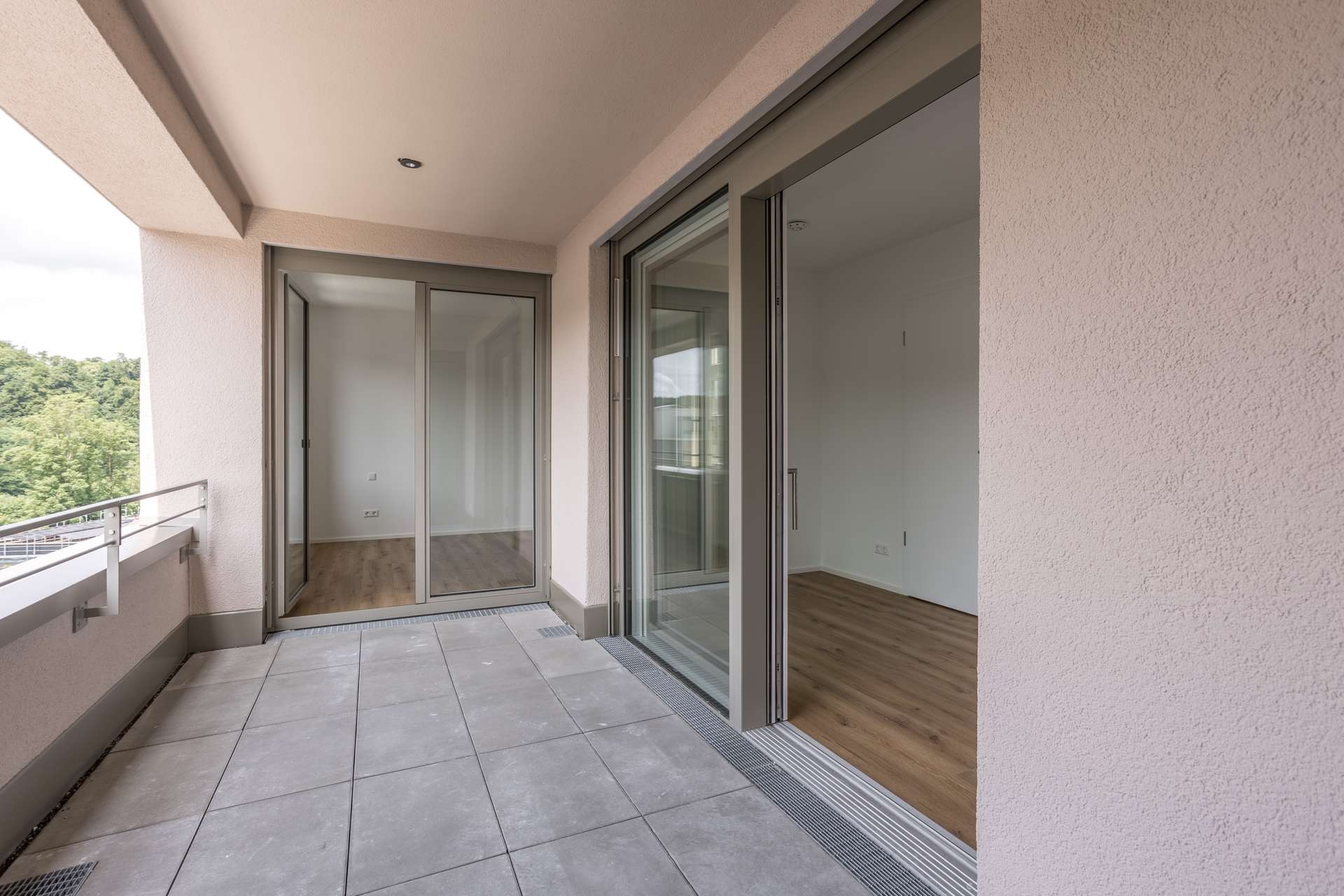 Ansicht Loggia Ein geräumiger Balkon mit hellgrauen Fliesen, Glasschiebetüren, die zu leeren Räumen mit Holzböden führen, und Blick ins Grüne. Der Bereich ist hell und modern.
