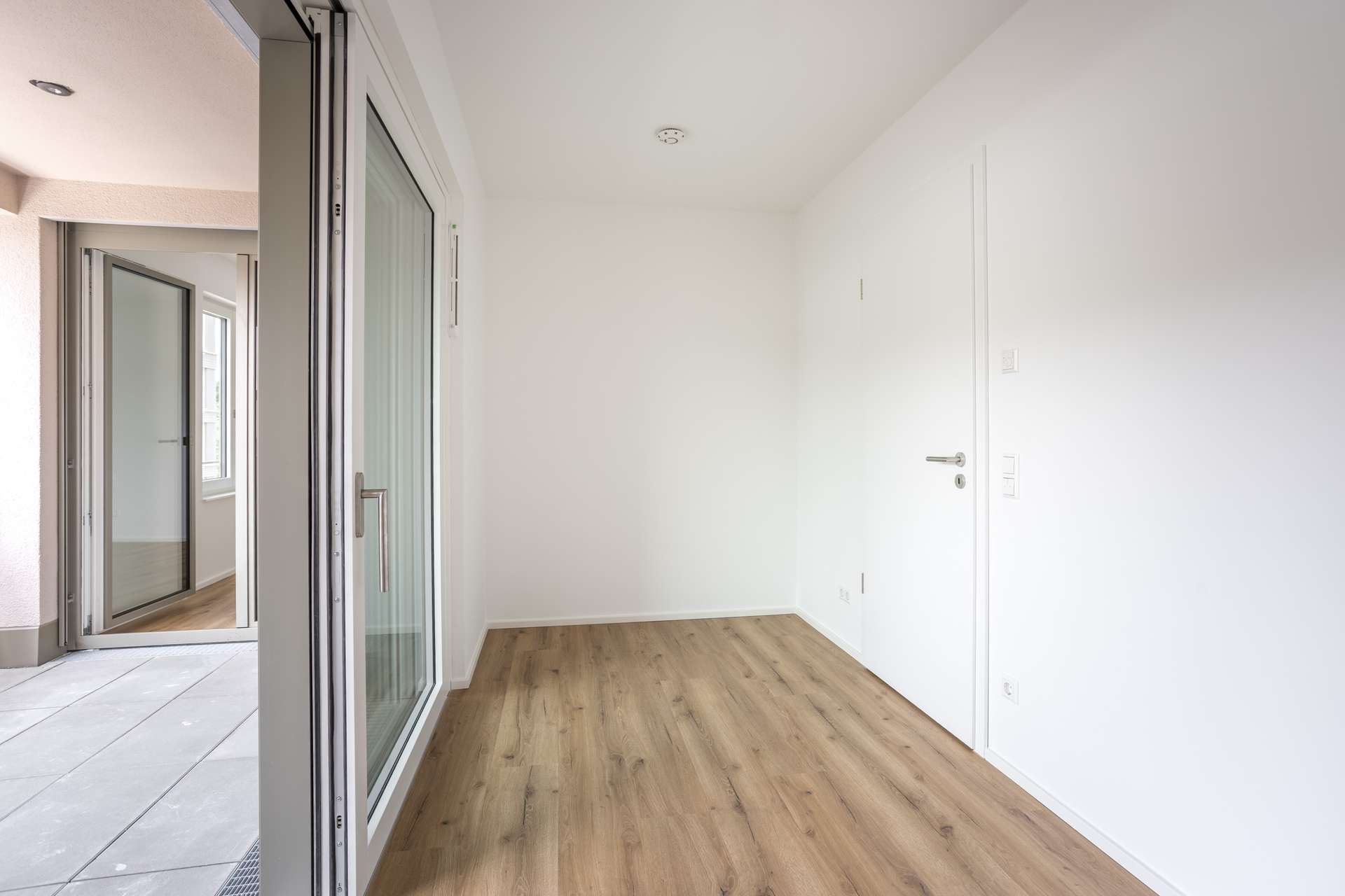 Ansicht Gast_Büro Ein heller, leerer Raum mit weißen Wänden, hellem Holzboden und großen Glasschiebetüren, die auf eine Außenterrasse führen. Der Raum ist von Tageslicht gut durchflutet.