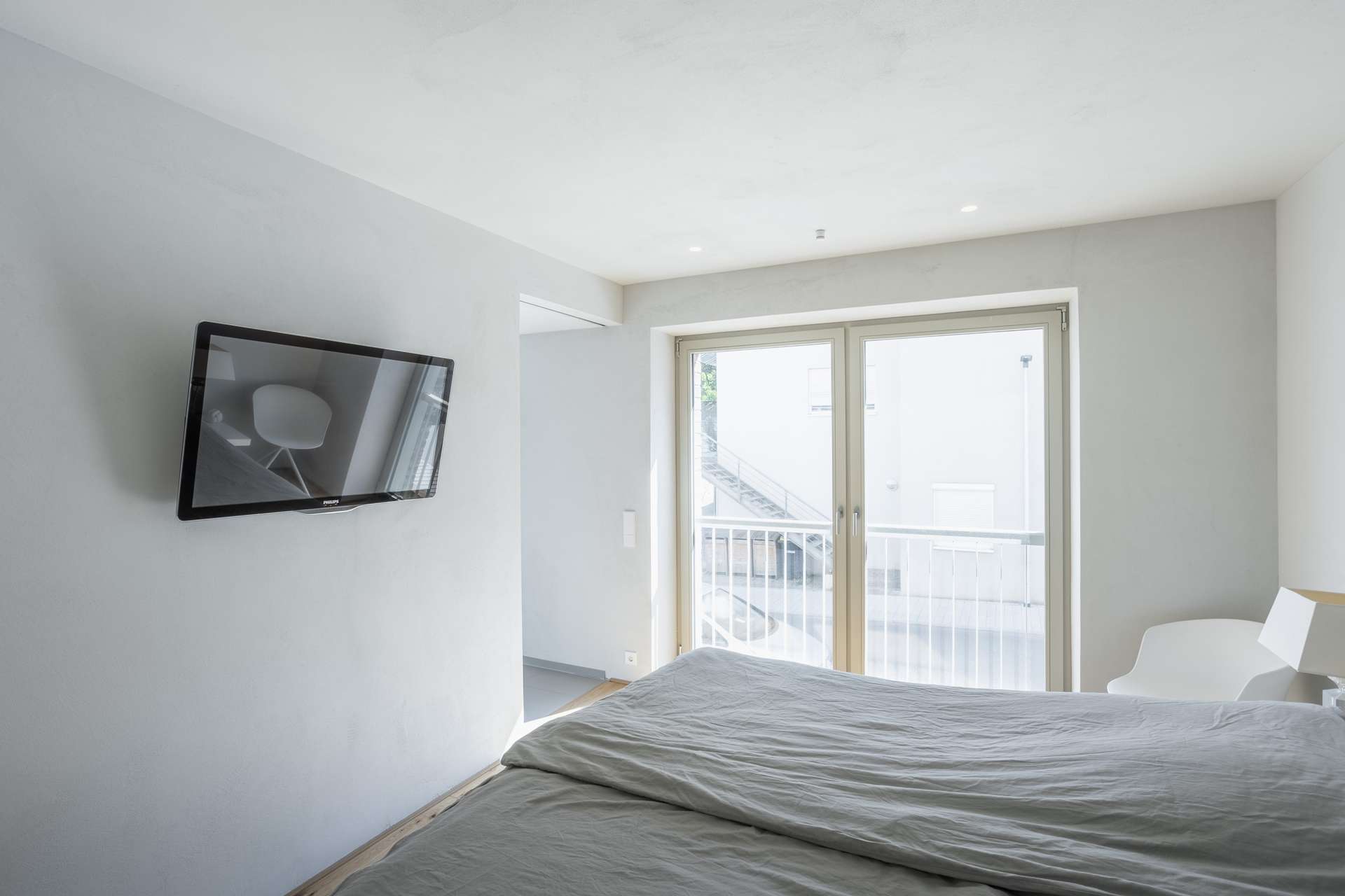 Schlafen Minimalistisch eingerichtetes Schlafzimmer mit einem an der Wand montierten Fernseher, einem ungemachten Bett, einem weißen Stuhl und großen Glastüren, durch die Tageslicht hereinströmt.