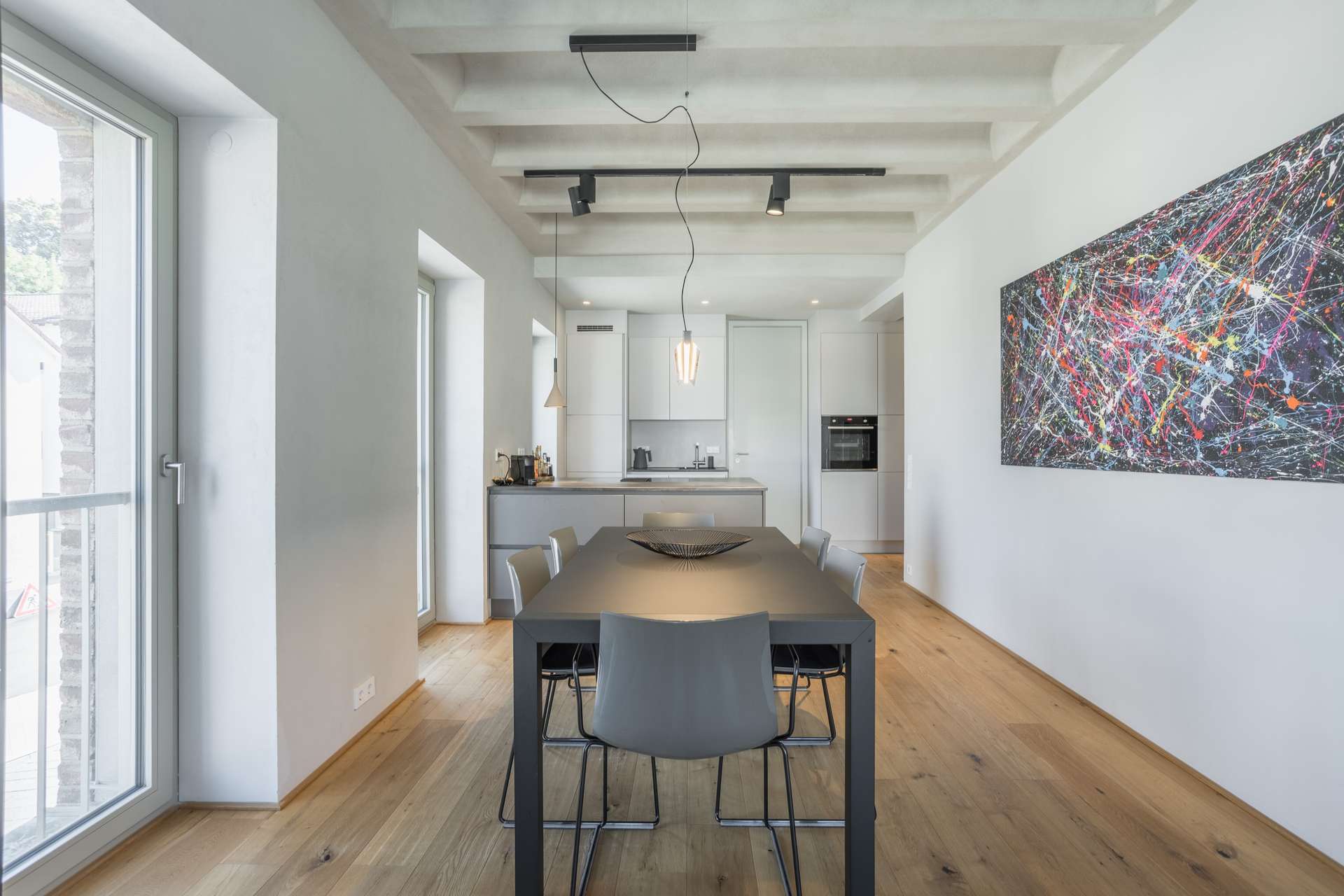 Essen Moderner Essbereich mit einem schwarzen Tisch, grauen Stühlen, Holzboden, abstrakter Wandkunst und Blick in eine minimalistische, weiße Küche. Große Fenster lassen Tageslicht herein.