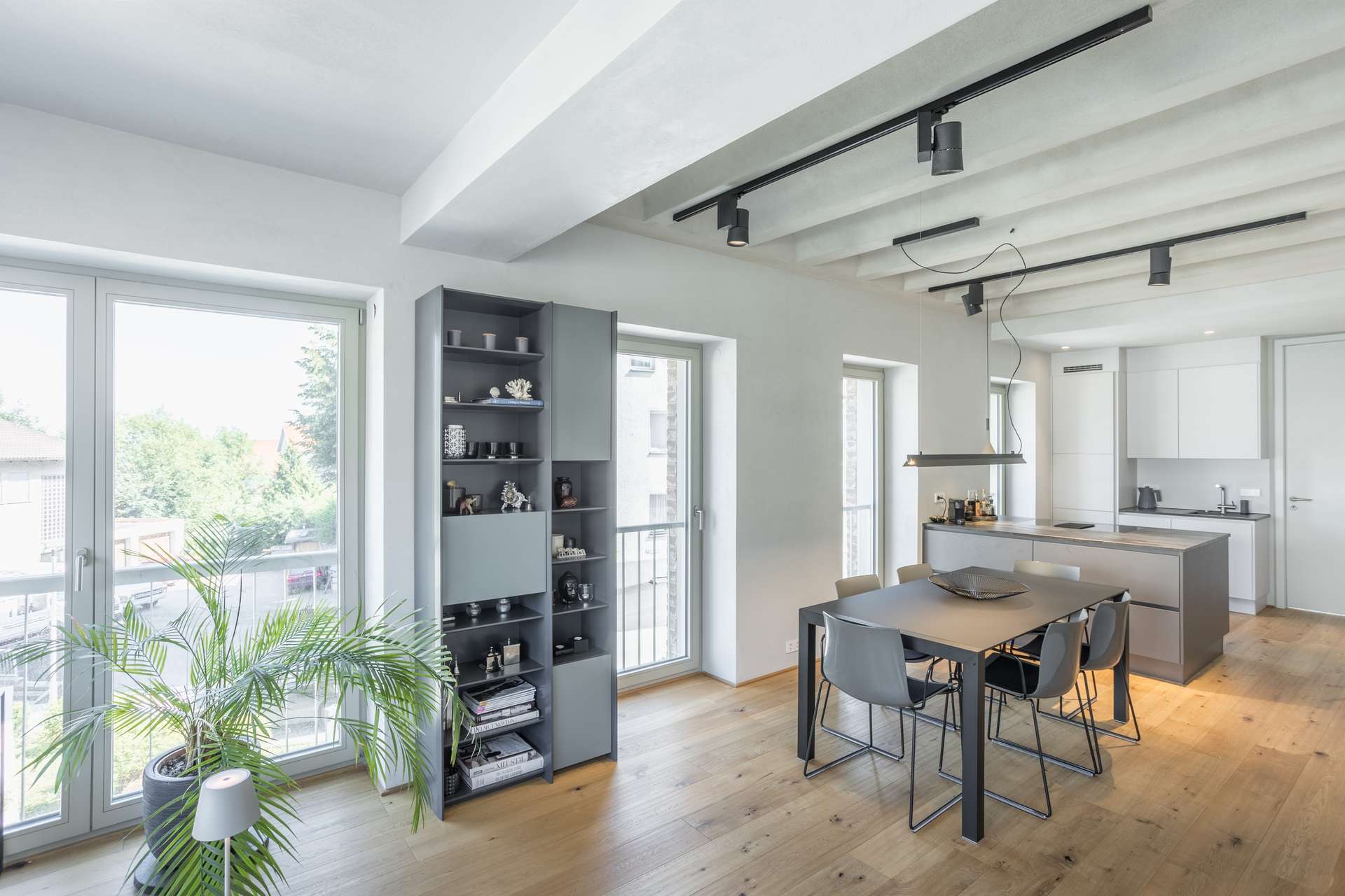 Küche/Essen Moderner Essbereich mit einem grauen Tisch und vier Stühlen, offenen Regalen mit Dekoartikeln, großen Fenstern, einer Topfpflanze und Blick in eine minimalistisch gestaltete Küche. Natürliches Licht durchflutet den Raum und bringt die Holzböden besonders gut zur Geltung.