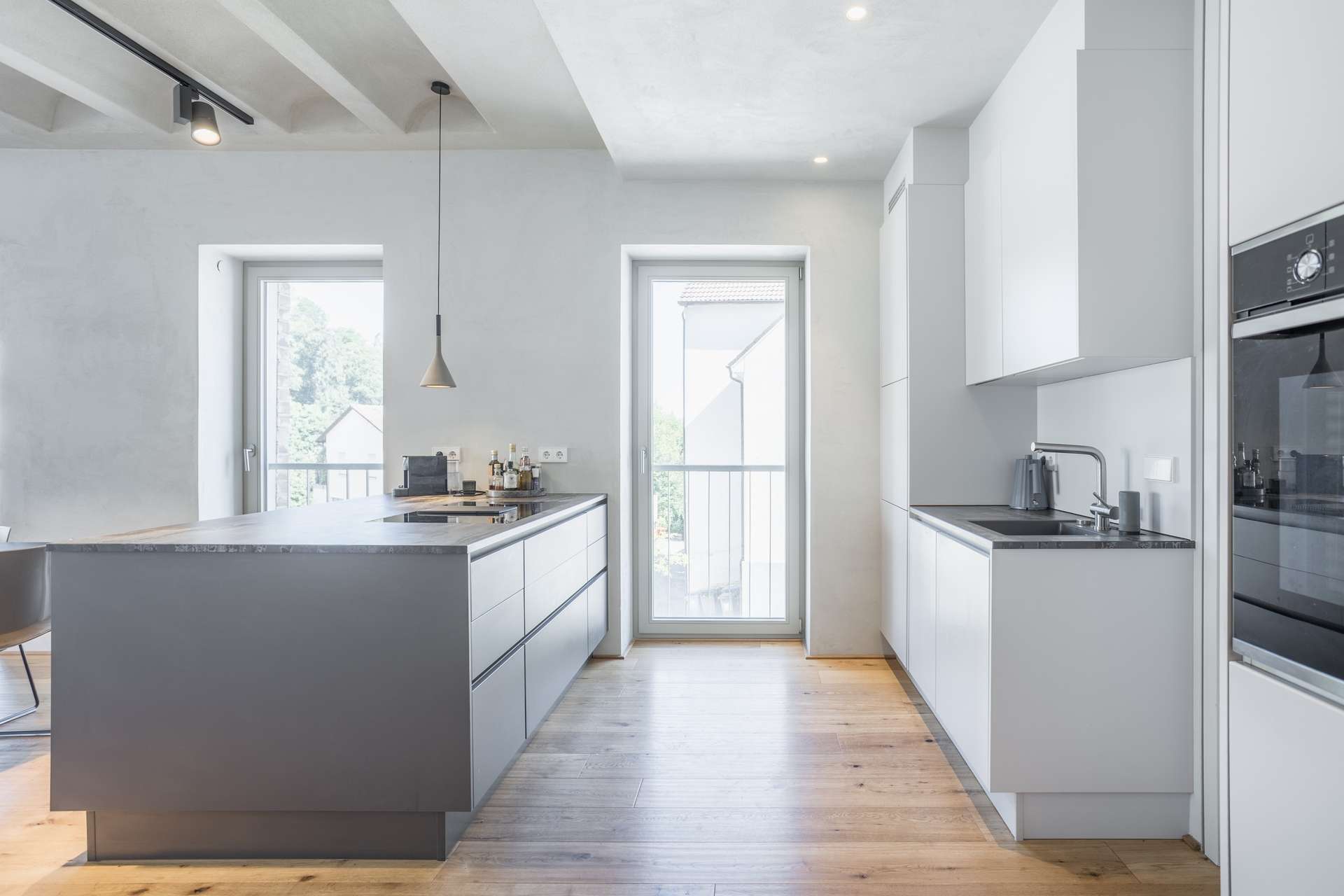 Küche Moderne Küche mit weißen Schränken, einer großen grauen Kücheninsel, Holzböden, Einbaubacköfen und zwei hohen Fenstern, durch die Tageslicht hereinströmt; minimalistisches und klares Design.