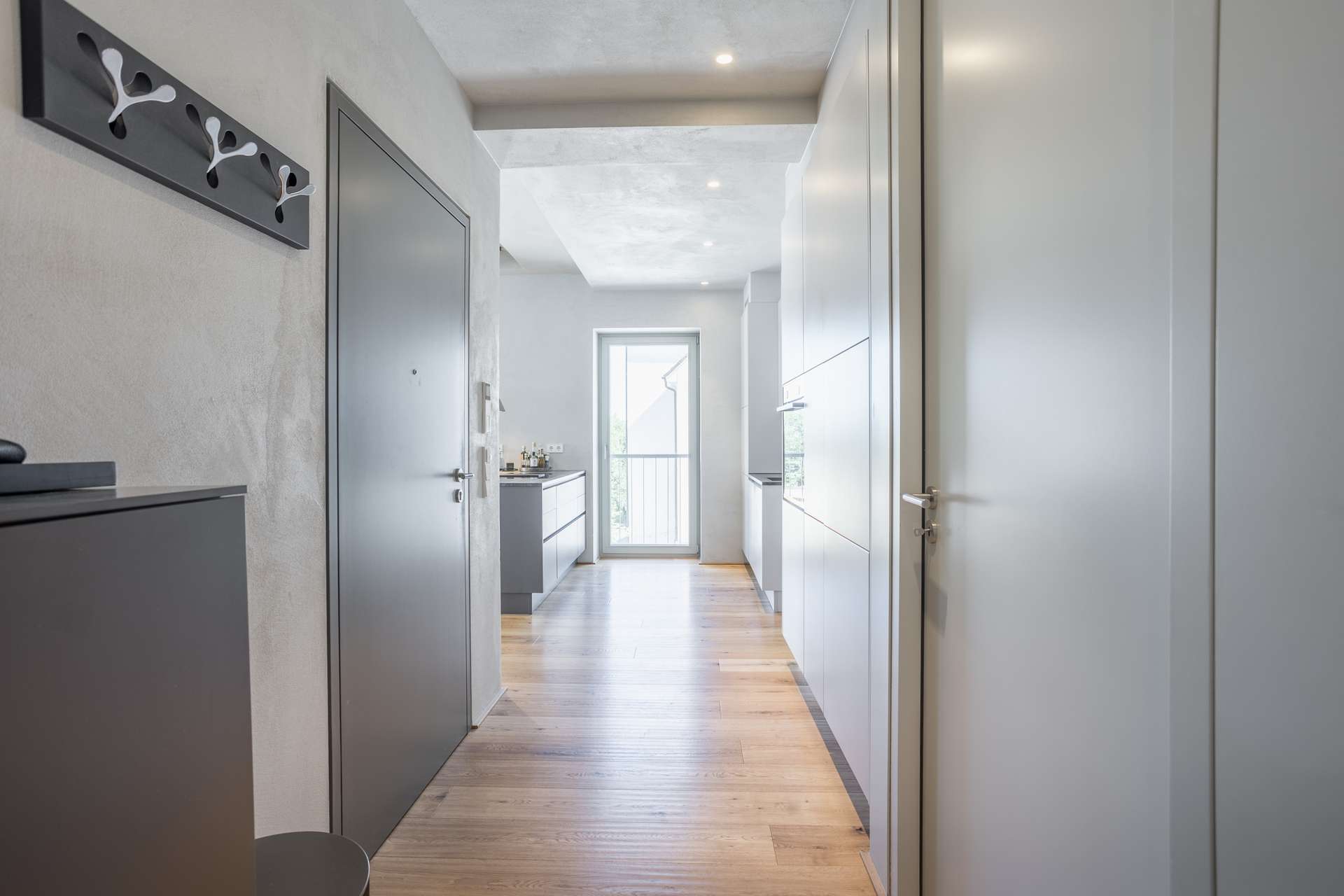 Flur Moderner Flur mit hellgrauen Wänden, Holzboden und minimalistischer Einrichtung. An der linken Wand ist eine Garderobe angebracht, und eine Tür am Ende führt in einen hellen Raum mit einem großen Fenster.
