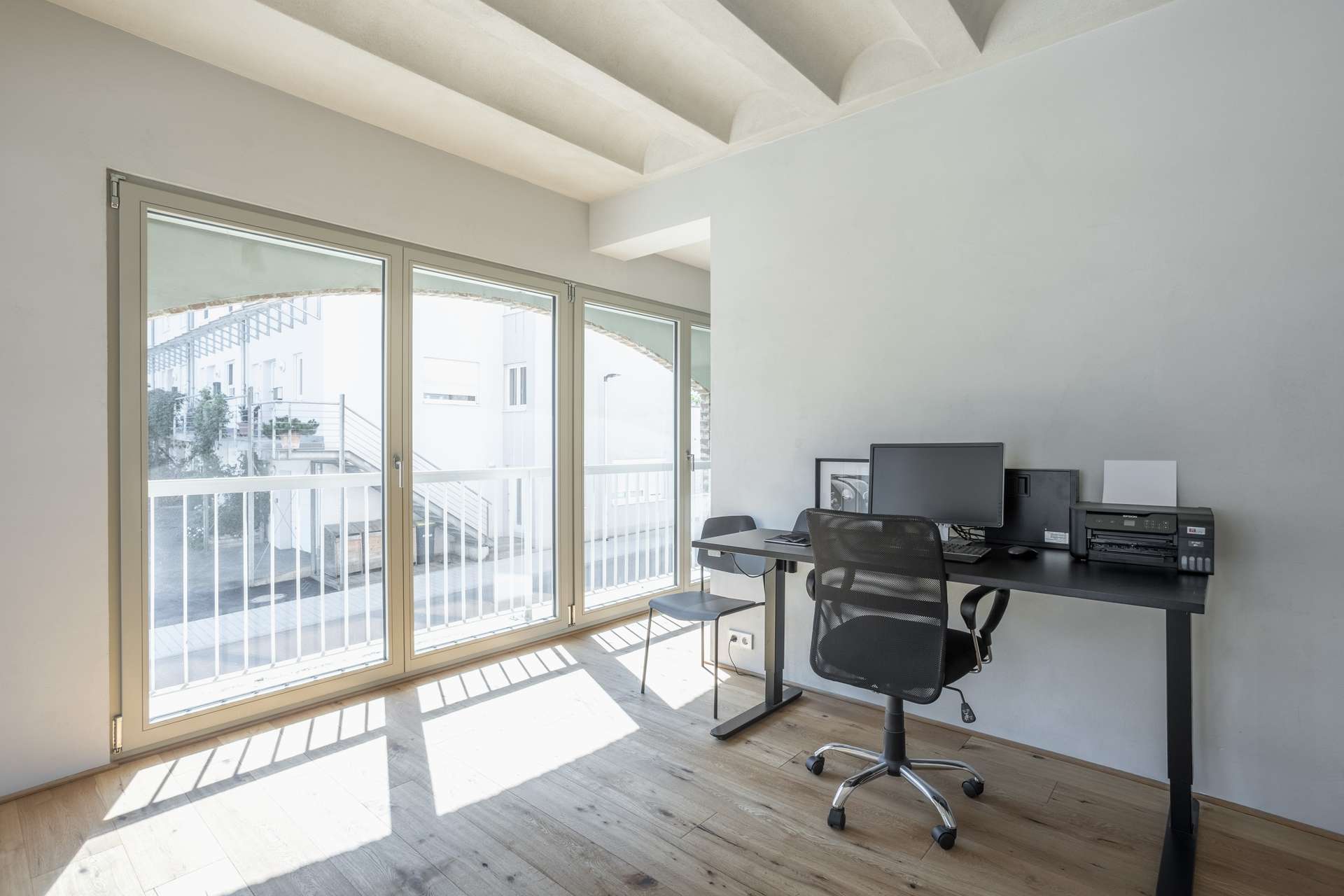 Büro Minimalistisches Homeoffice mit großen Fenstern, einem schwarzen Schreibtisch, einem Bürostuhl, einem Computer, einem Drucker und einem Stuhl. Der Raum wird von Tageslicht durchflutet, und durch die Fenster hat man einen Blick auf eine Treppe und den Außenbereich.