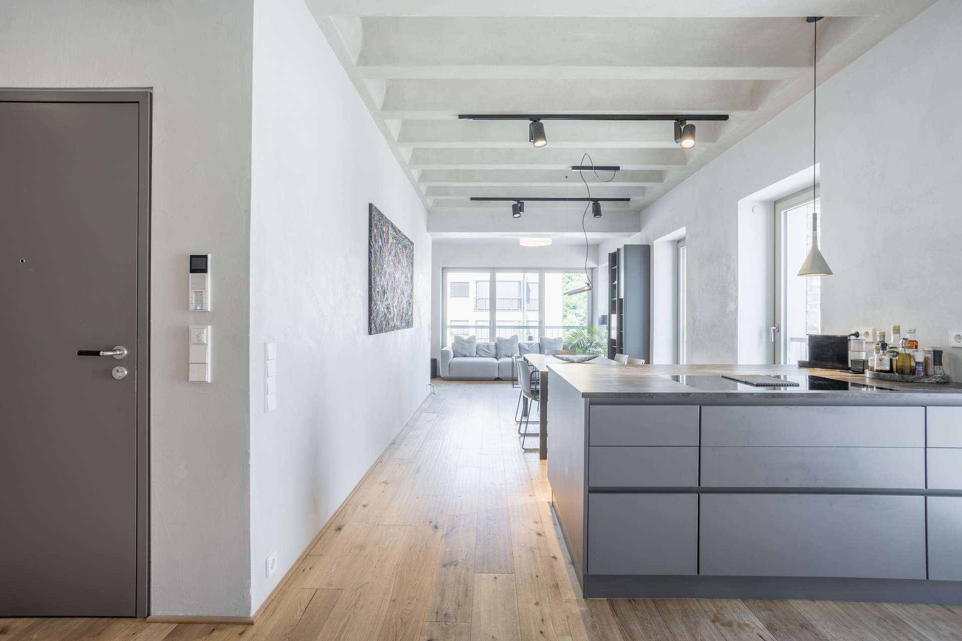 Küche/Wohnen/Essen Eine moderne, minimalistische Küche mit grauen Schränken und einem Holzboden, der in ein helles Wohnzimmer mit einem Sofa und großen Fenstern im Hintergrund übergeht.