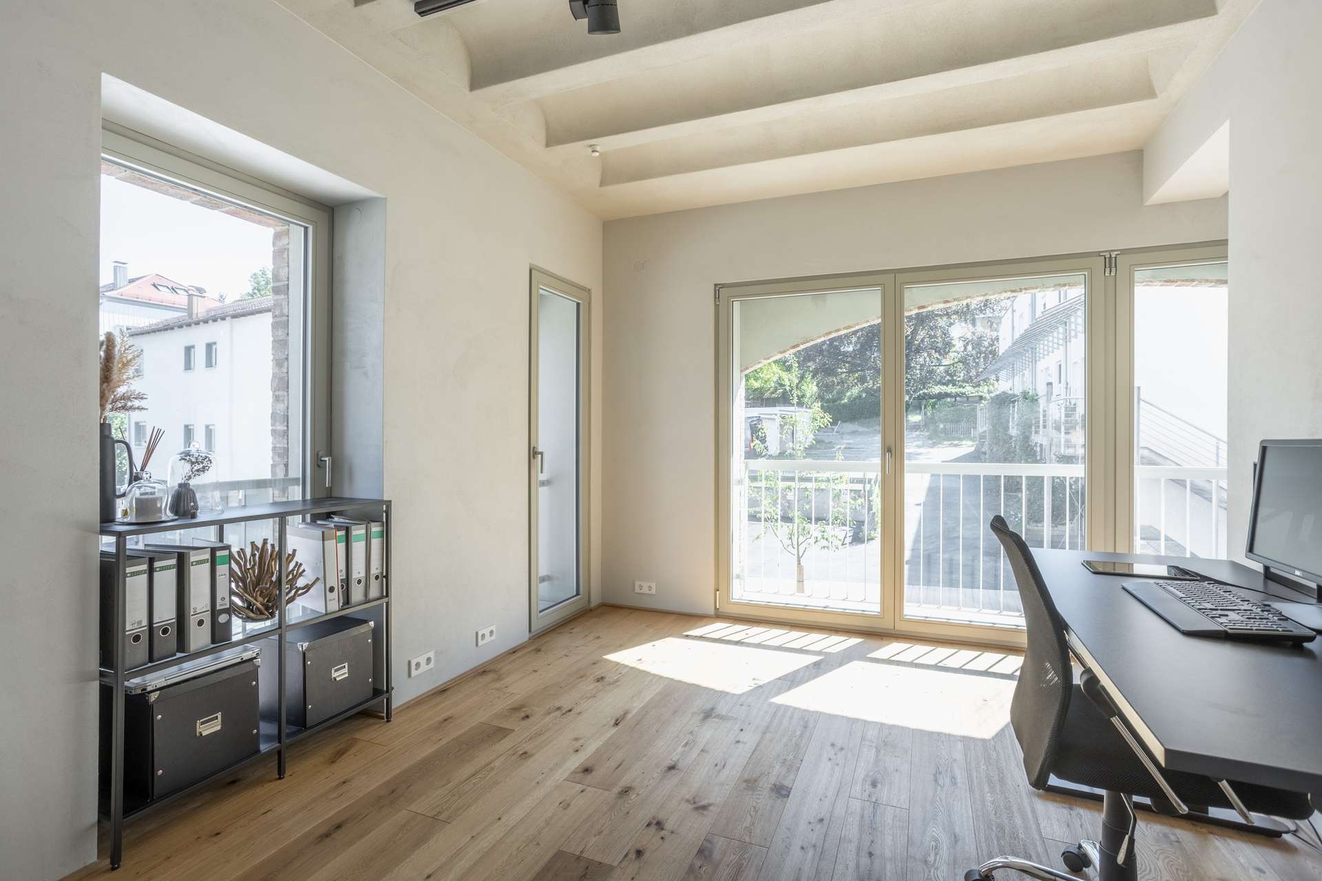 Büro Ein helles, modernes Büro mit Holzboden, großen Fenstern, einem Schreibtisch mit Computer, einem schwarzen Stuhl und einem Regal mit Ordnern und Dekoartikeln. Sonnenlicht durchflutet den minimalistisch gestalteten Raum.