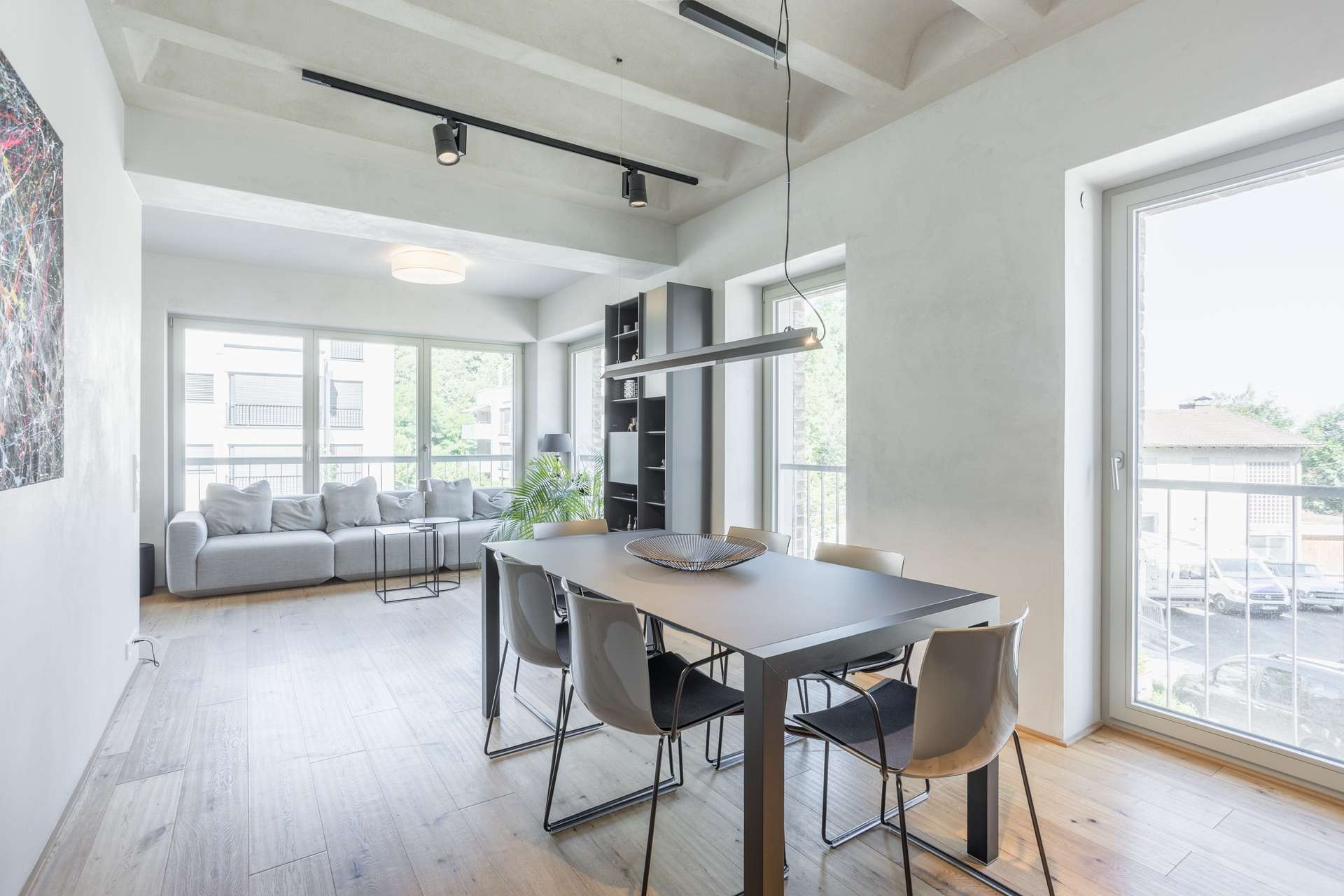 Wohnen/Essen Moderner Essbereich mit einem schwarzen Tisch und vier Stühlen neben großen Fenstern. Im Hintergrund sind ein graues Sofa und Bücherregale zu sehen; reichlich Tageslicht durchflutet den hellen, minimalistischen Raum.