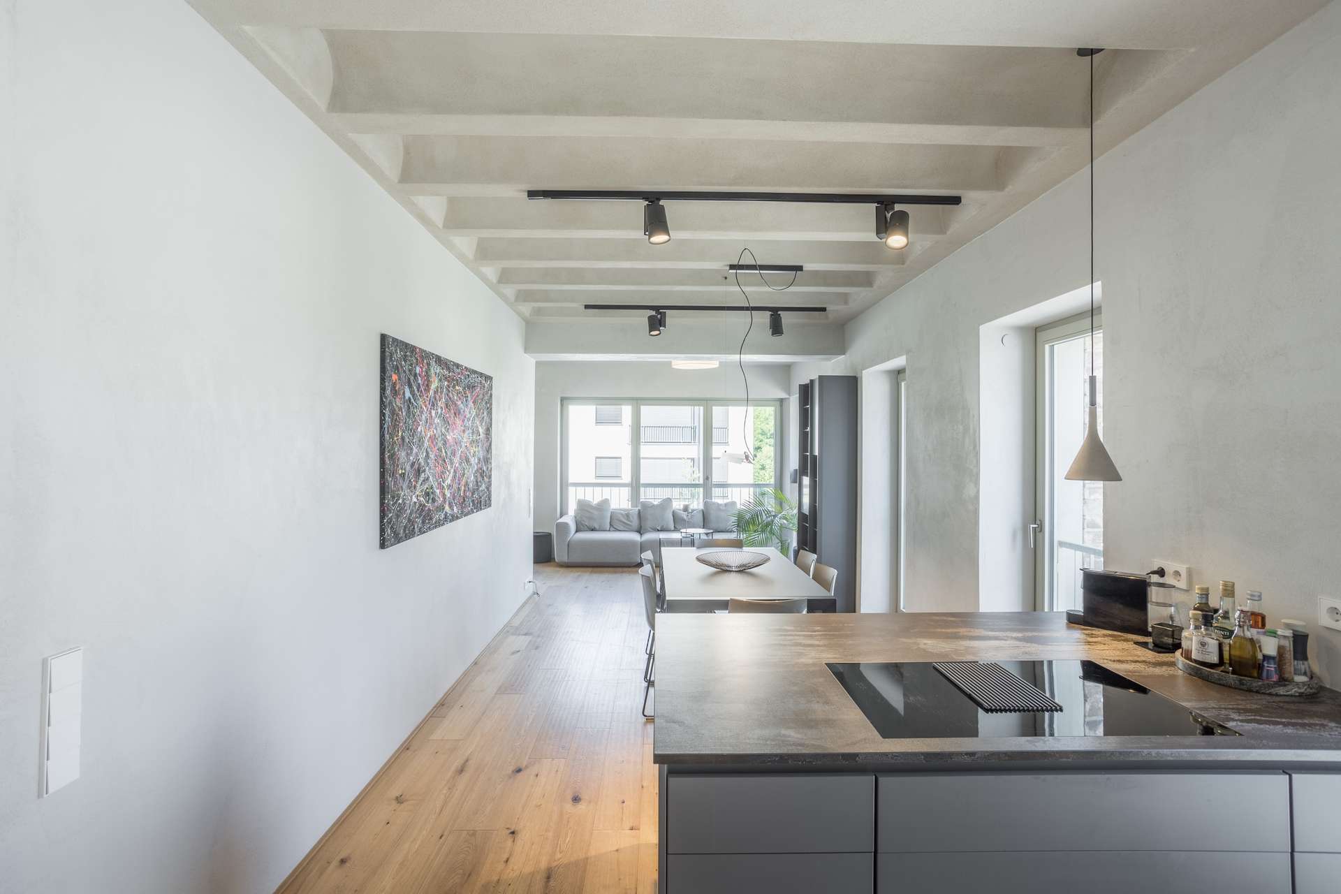 Küche/Wohnen/Essen Moderne, minimalistische Küche mit einer grauen Kücheninsel und einem grauen Kochfeld, offen zu einem hellen Wohnzimmer mit einem grauen Sofa, abstrakten Wandbildern und großen Fenstern, durch die Tageslicht hereinströmt.
