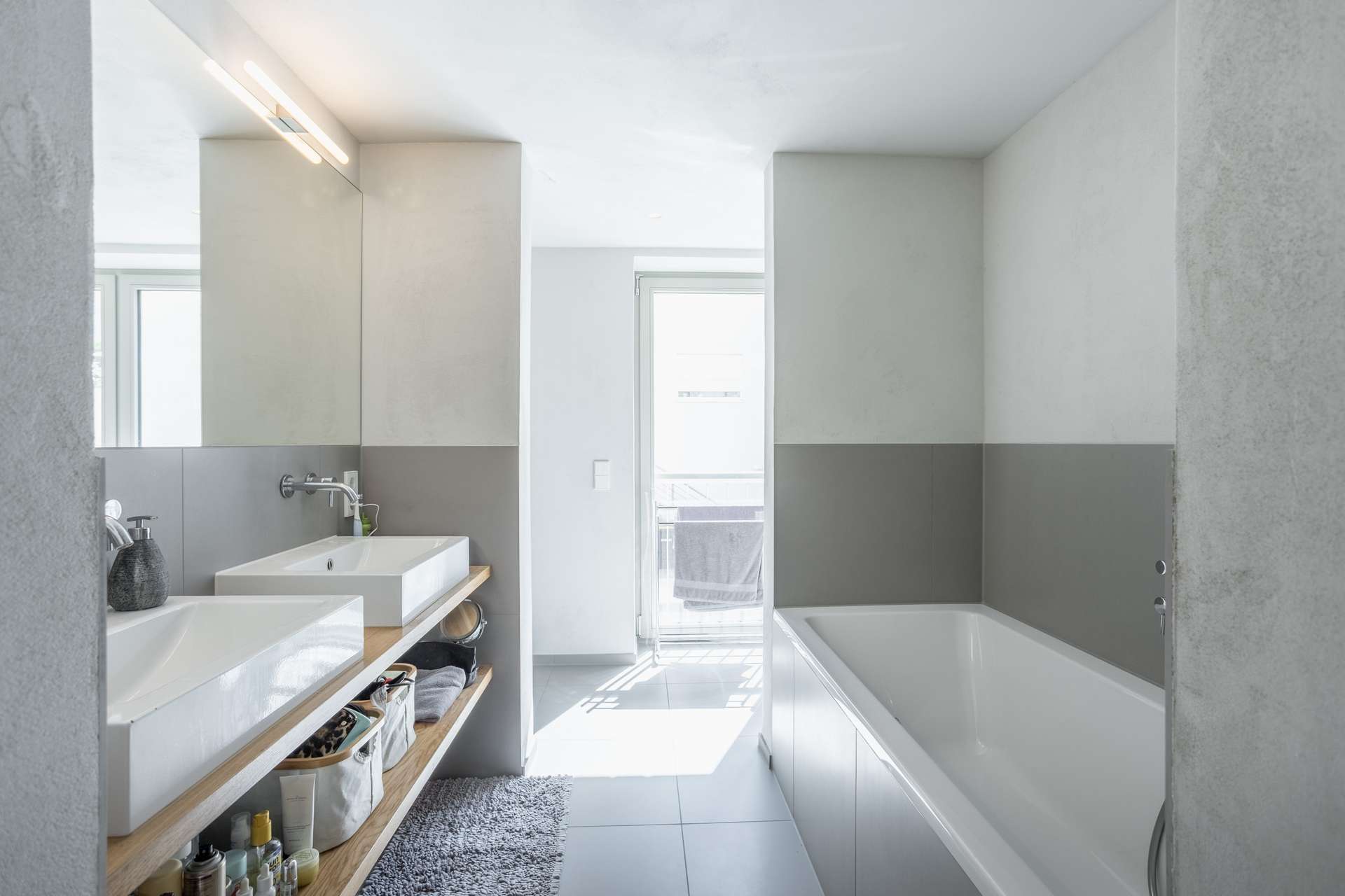 Bad Modernes Badezimmer mit einer weißen Badewanne, einem großen Spiegel, zwei Waschbecken auf einem Holzregal, Aufbewahrungskörben darunter und einem Fenster, durch das Tageslicht hereinfällt. Grau- und Weißtöne sorgen für einen klaren, minimalistischen Look.