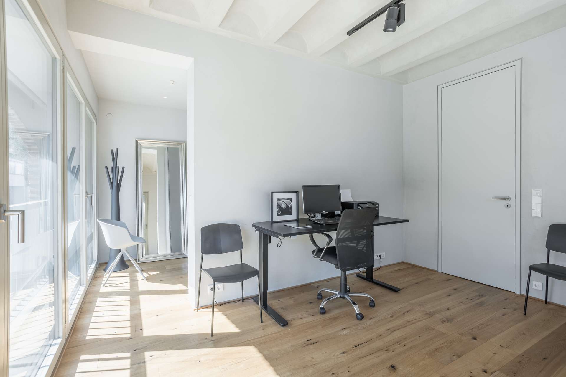 Büro Minimalistisches Homeoffice mit einem Schreibtisch, einem Computer, einem Bürostuhl und zwei schwarzen Stühlen. Durch große Glastüren fällt Sonnenlicht herein. Im Hintergrund sind ein kleines gerahmtes Bild und eine moderne Garderobe zu sehen.