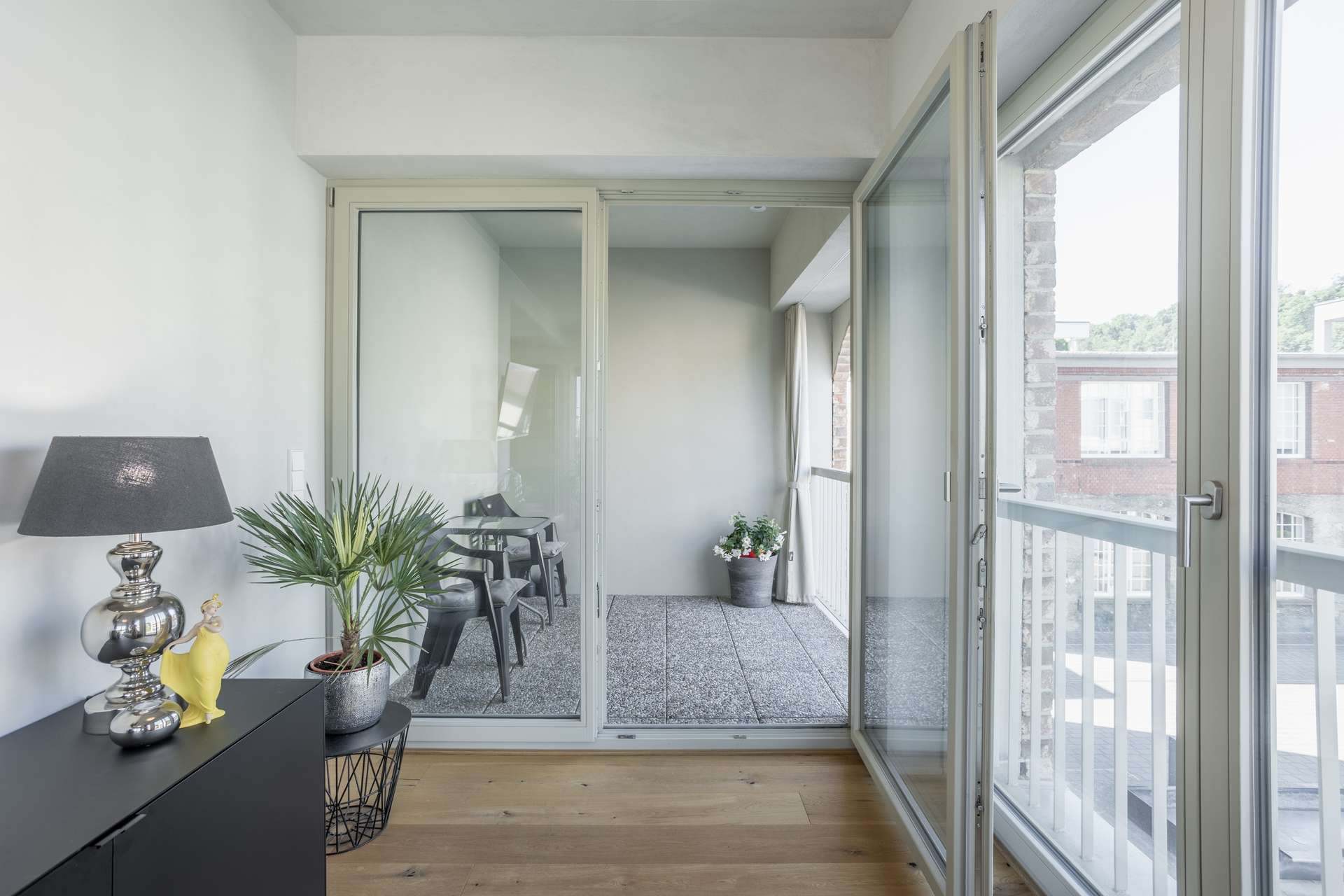 Loggia Ein heller Raum mit Glastüren, die auf einen Balkon führen. Im Raum stehen ein schwarzer Schrank mit einer Lampe, eine kleine Pflanze und eine gelbe Figur. Draußen stehen zwei Stühle, ein Tisch und eine Topfpflanze.