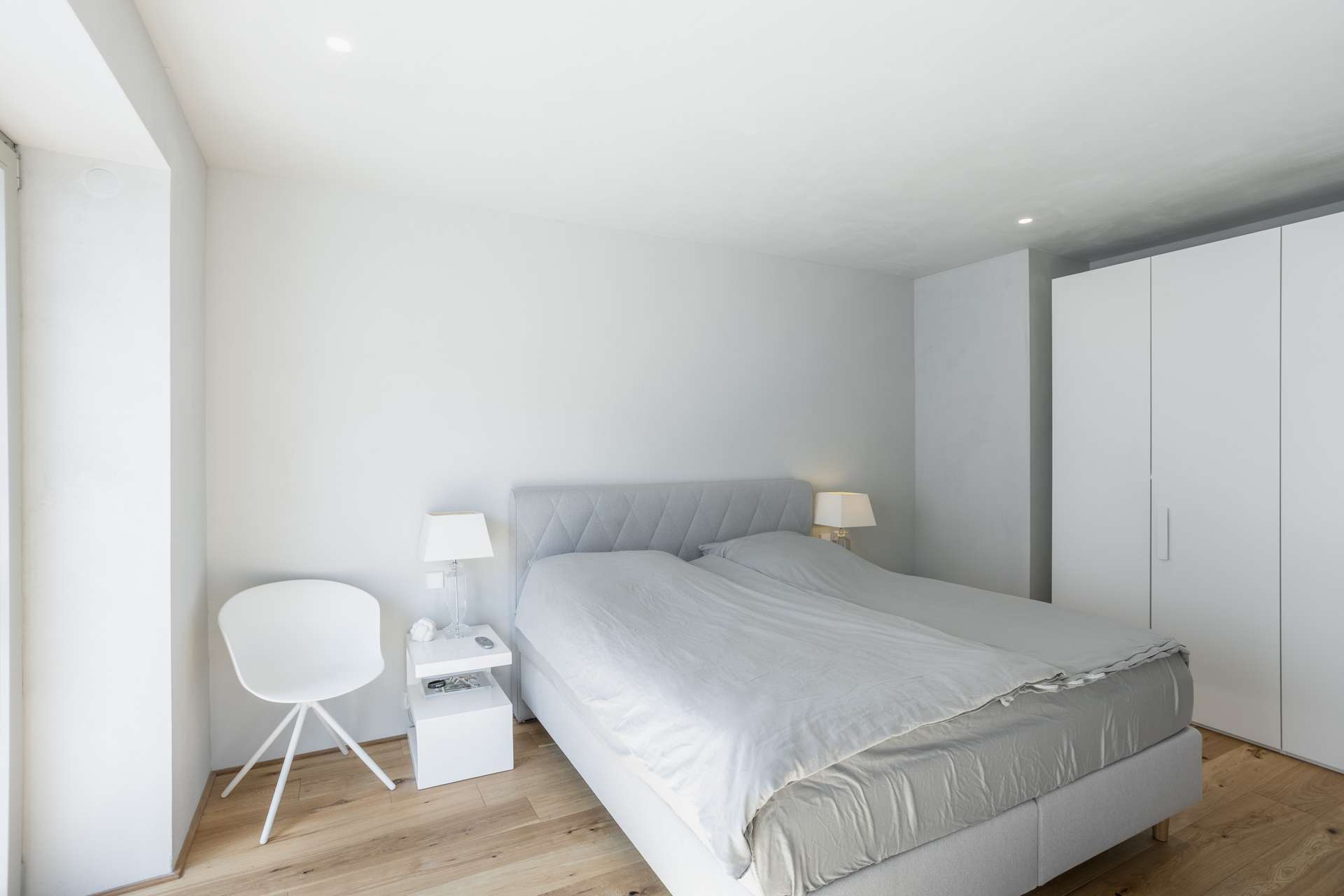 Schlafen Ein minimalistisches Schlafzimmer mit einem Doppelbett, zwei Nachttischen mit Lampen, einem weißen Stuhl und einem großen weißen Kleiderschrank. Der Raum hat helle Wände, einen Holzboden und erhält Tageslicht durch ein Fenster auf der linken Seite.