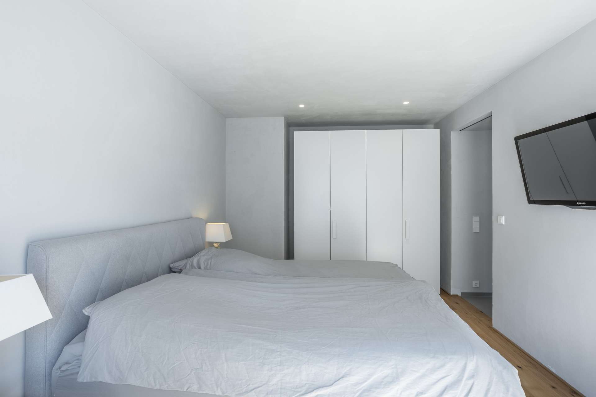 Schlafen Minimalistisches Schlafzimmer mit einem Doppelbett, hellgrauer Bettwäsche, zwei weißen Nachttischen mit Lampen, einem weißen Kleiderschrank, Holzboden und einem an der Wand montierten Fernseher. Der Raum besticht durch ein klares, modernes Design in neutralen Farben.