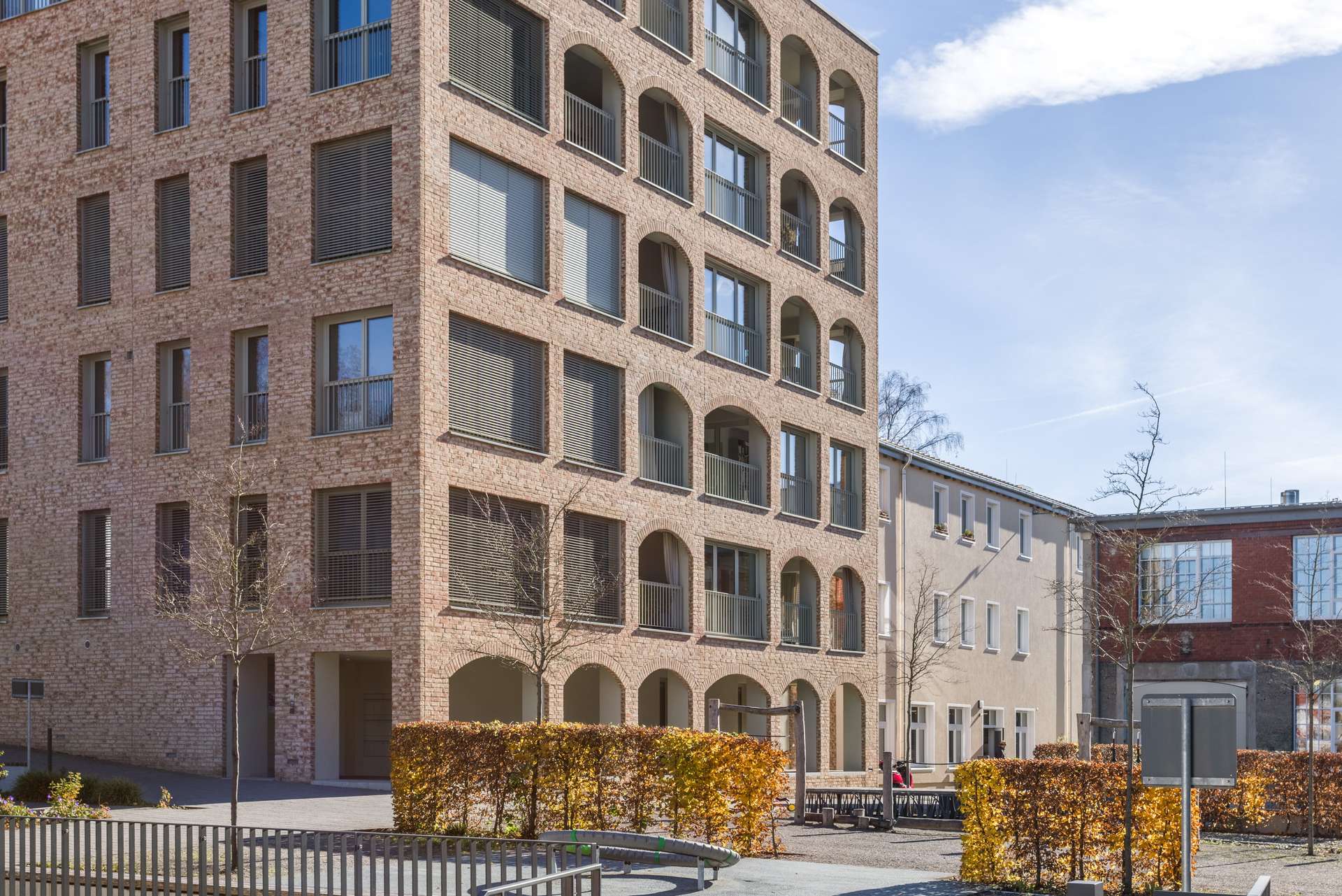 Außenansicht Modernes Mehrfamilienhaus mit großen Bogenfenstern und Backsteinfassade, direkt neben einem von Hecken gesäumten Gehweg und einem kleinen Innenhof. Der blaue Himmel und die kahlen Bäume lassen auf den frühen Frühling oder den späten Herbst schließen.