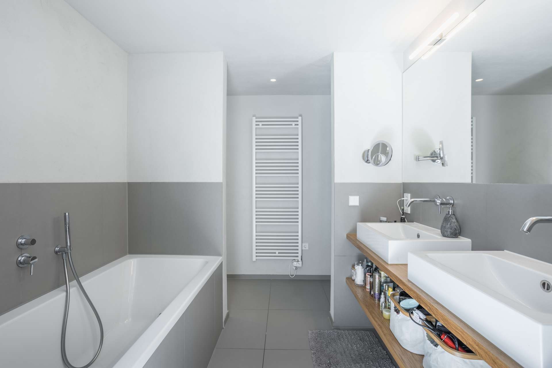Bad Modernes Badezimmer mit einer Badewanne auf der linken Seite, einem Doppelwaschbecken auf einem Waschtisch aus Holz mit Ablagen auf der rechten Seite, einem Wandspiegel und einem weißen Handtuchheizkörper an der Rückwand. Das Design ist minimalistisch und in neutralen Farben gehalten.