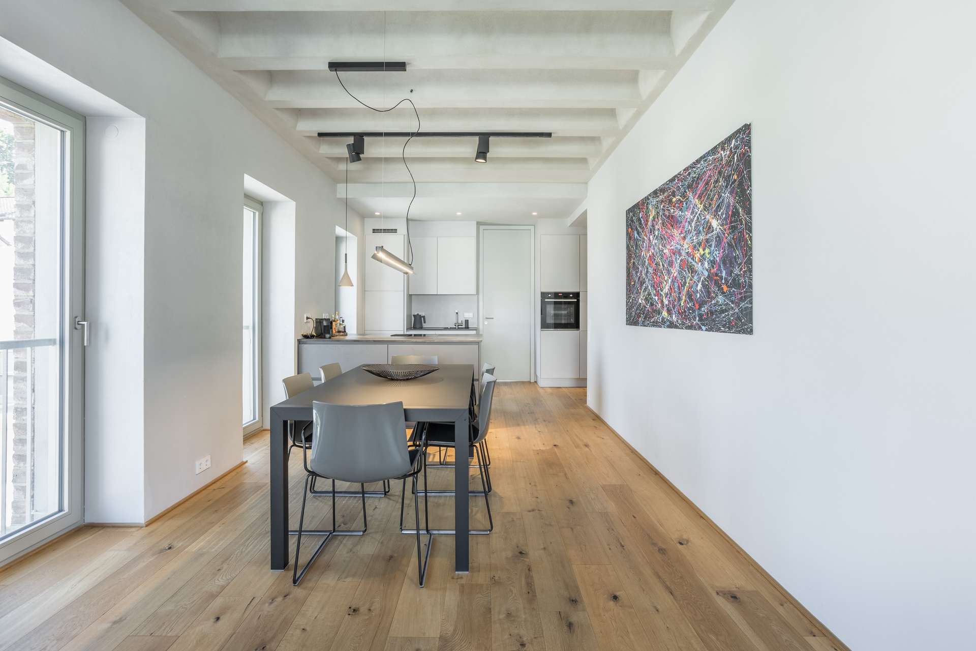 Essen Modernes Esszimmer mit hellen Holzböden, grauem Tisch und Stühlen, zeitgemäßer Beleuchtung, abstrakten Wandbildern und großen Fenstern, durch die Tageslicht hereinströmt; im Hintergrund schließt es an eine offene Küche an.
