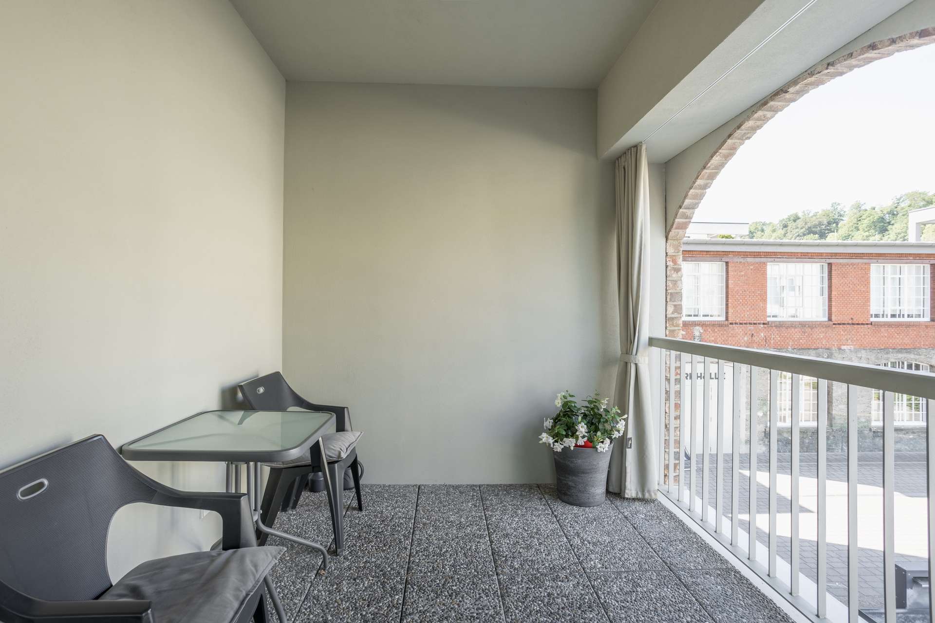 Loggia Ein kleiner Balkon mit zwei schwarzen Stühlen, einem Tisch mit Glasplatte, einer Topfpflanze und einem weißen Geländer. Durch eine gewölbte Öffnung sind draußen ein Backsteingebäude und Grünflächen zu sehen.