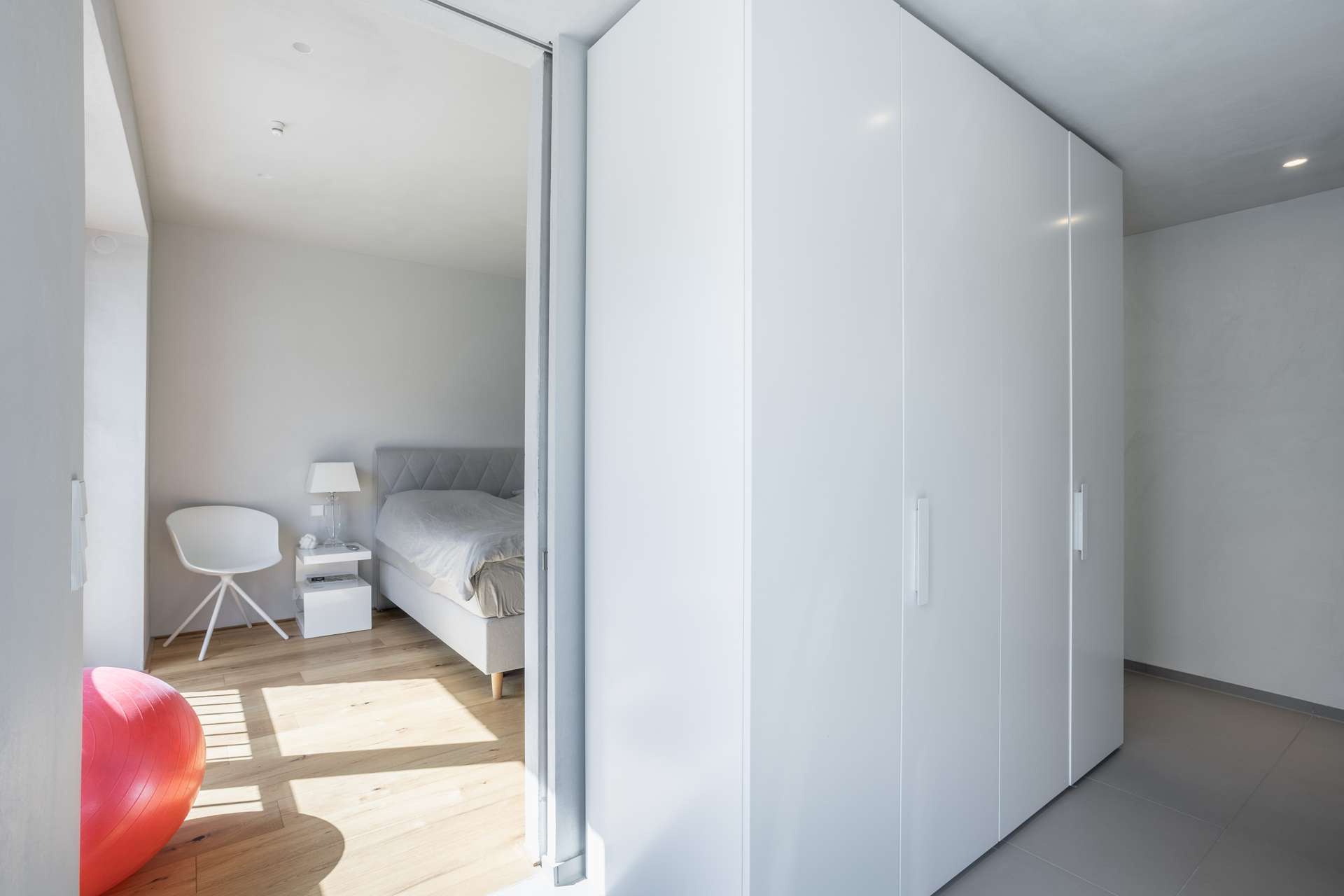 Schlafen Ein modernes Schlafzimmer mit einem weißen Bett, einem Nachttisch, einem Stuhl und einer Lampe. Rechts stehen große weiße Kleiderschränke. Auf dem Holzboden neben einem Fenster, durch das Sonnenlicht hereinströmt, liegt ein roter Gymnastikball.
