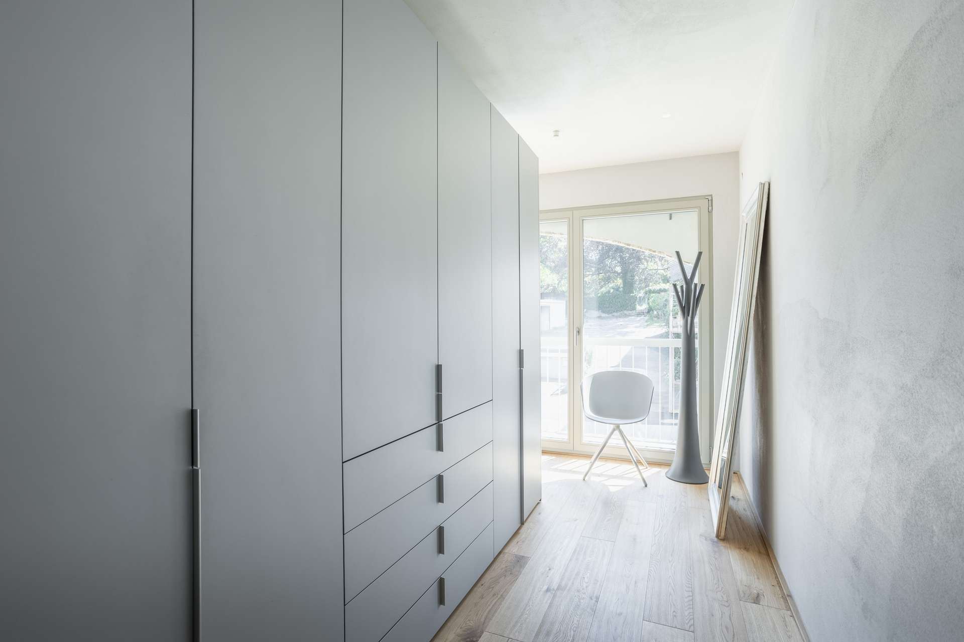 Ankleide Ein minimalistisch gestalteter Kleiderschrankbereich mit großen grauen Kleiderschränken, hellem Holzboden, einem weißen Stuhl, einer Garderobe und einem großen Fenster, durch das Tageslicht hereinströmt und eine helle und klare Atmosphäre schafft.