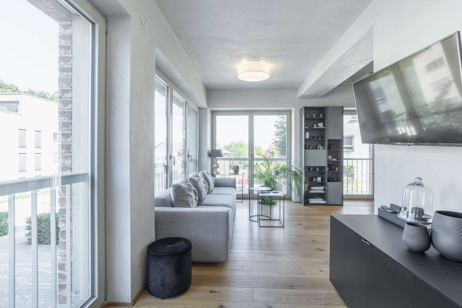 Wohnen Modernes Wohnzimmer mit großen Fenstern, einem grauen Sofa, einem Couchtisch aus Glas, einem an der Wand montierten Fernseher, offenen Regalen, hellem Holzboden und minimalistischer Einrichtung. Der Raum wird von natürlichem Licht durchflutet.