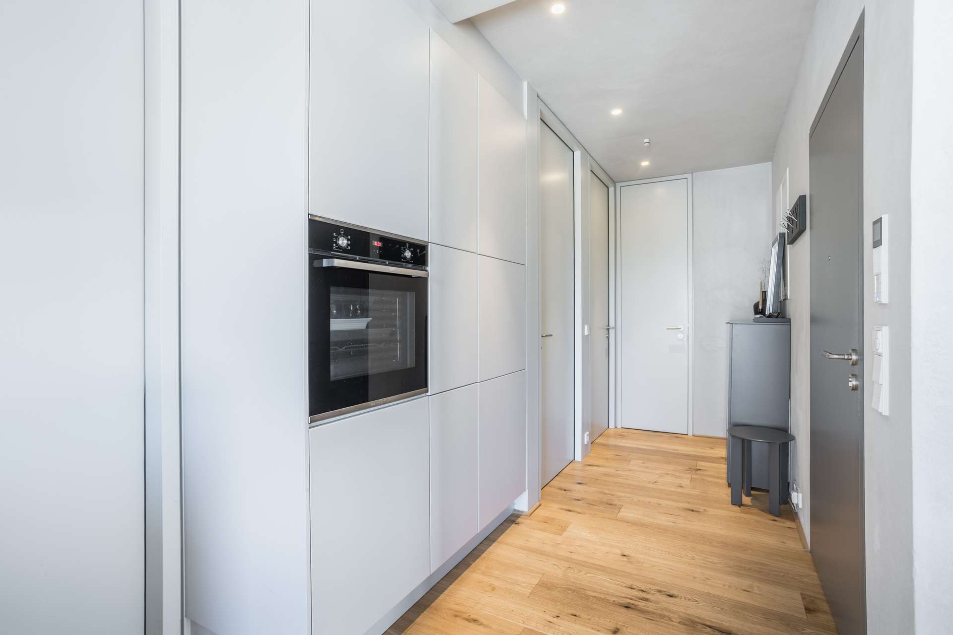 Flur Moderner Flur mit hellgrauen Einbauschränken, einem Einbaubackofen, Holzboden und Einbauleuchten an der Decke. Auf der rechten Seite, fast am Ende des Flurs, sind eine graue Tür und ein kleiner Tisch mit einigen Gegenständen zu sehen.