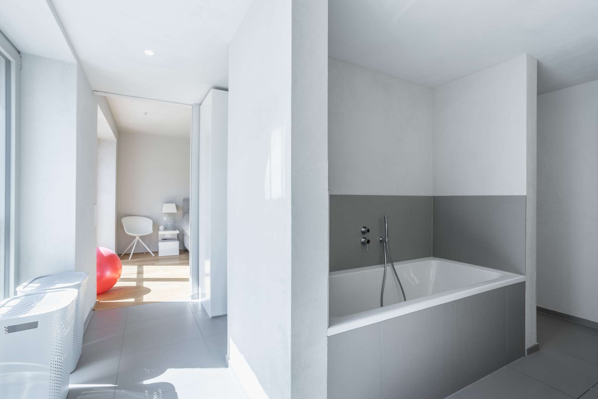 Bad Modernes Badezimmer in Grau- und Weißtönen mit einer rechteckigen Badewanne. Durch eine offene Tür ist ein angrenzender Raum mit einem Stuhl, einer Lampe und einem roten Gymnastikball zu sehen. Helles Tageslicht durchflutet den Raum.