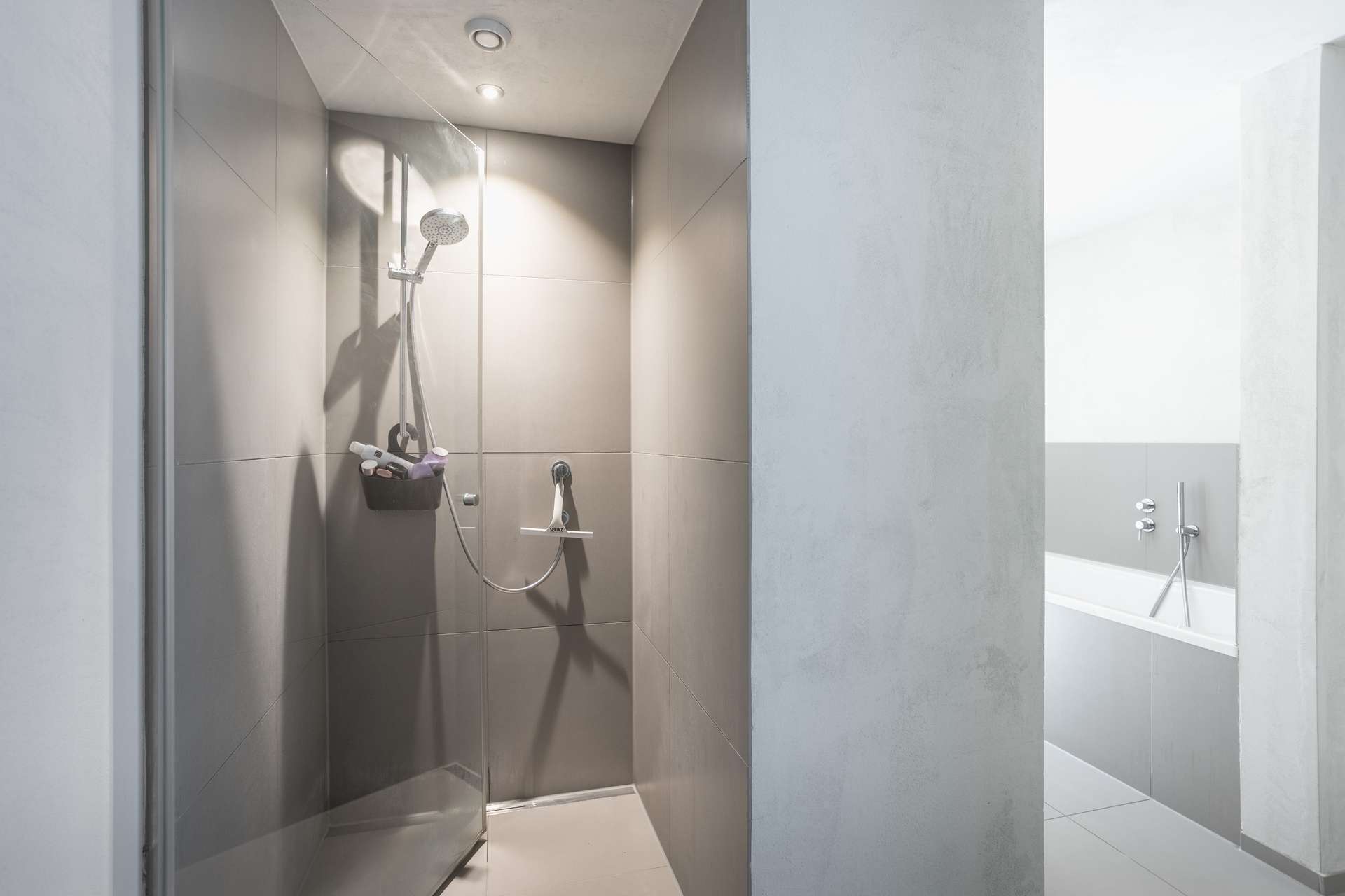 Bad Minimalistisches Badezimmer mit einer mit grauen Fliesen ausgekleideten begehbaren Dusche, einer Glastür und einem an der Decke montierten Duschkopf. Im Hintergrund ist in einem separaten Bereich eine weiße Badewanne zu sehen. Helles, klares und modernes Design.