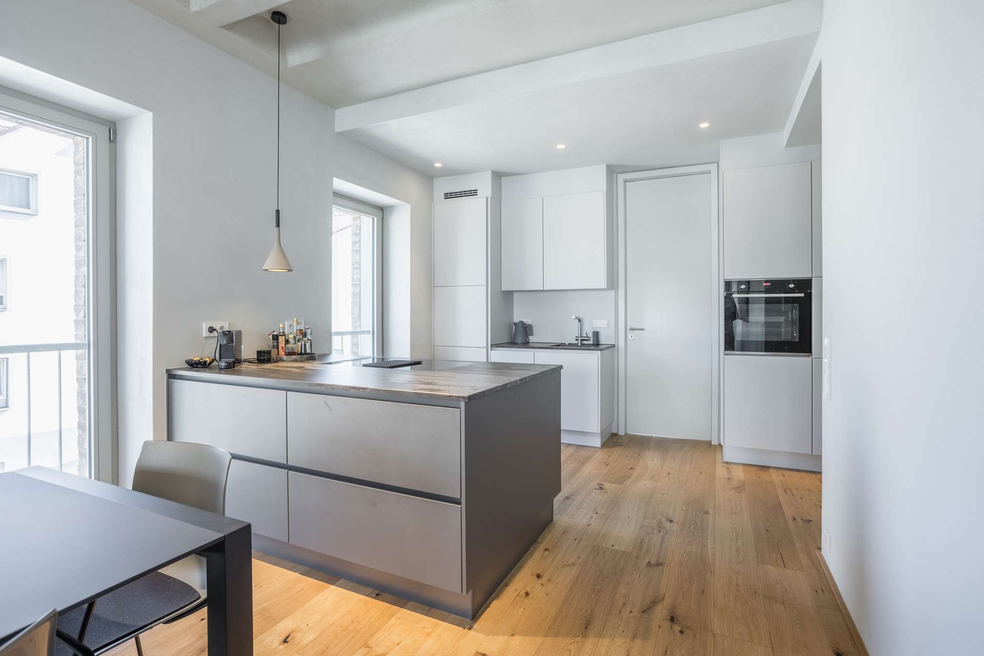 Küche Moderne Küche mit weißen Schränken, Holzboden, einer zentralen Kücheninsel mit Holzarbeitsplatte, Einbaugeräten, einer Pendelleuchte und großen Fenstern, durch die Tageslicht hereinströmt. Im Vordergrund ist teilweise ein kleiner Esstisch zu sehen.