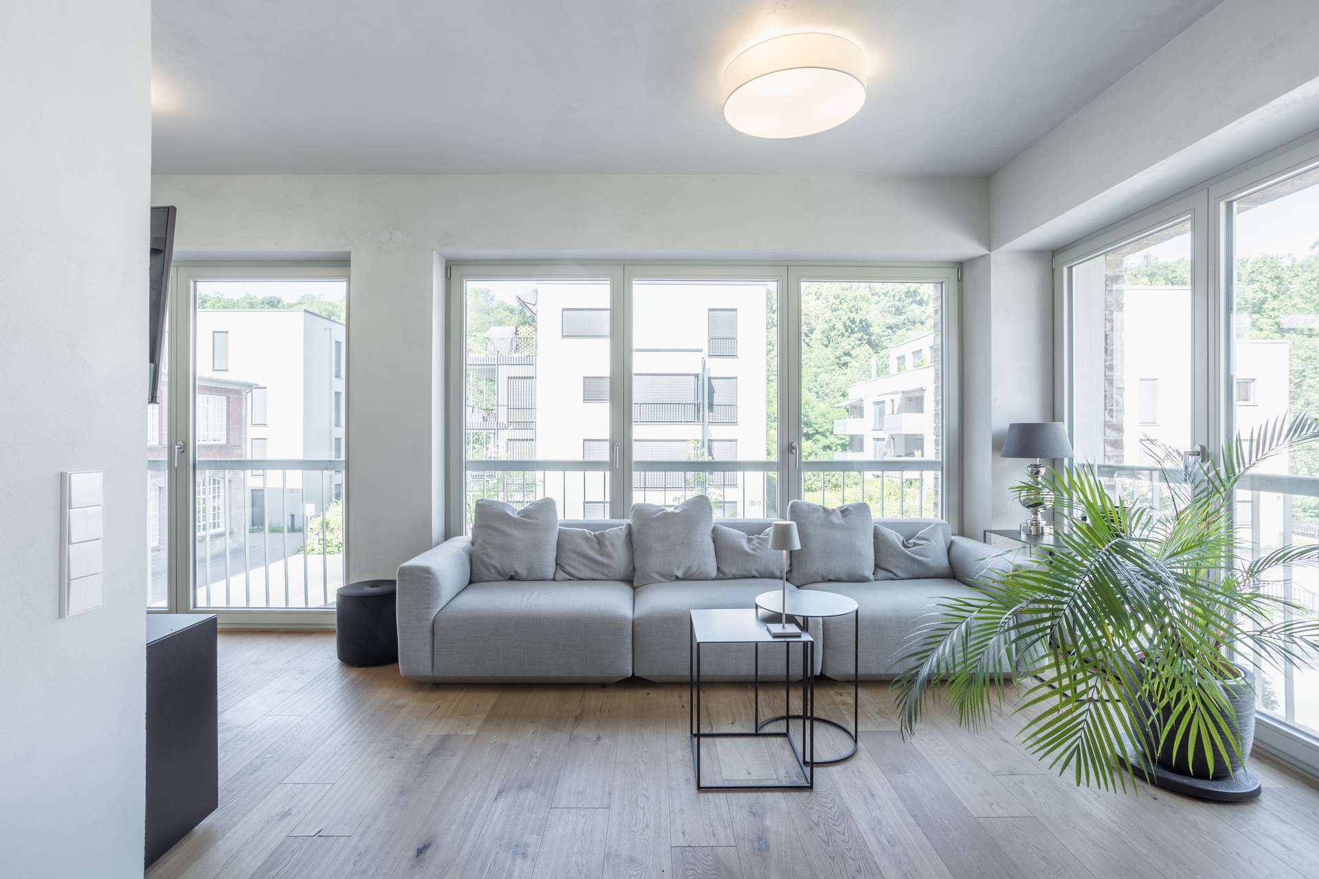 Wohnen Helles, modernes Wohnzimmer mit großen Fenstern, einem hellgrauen Sofa mit Kissen, einer Zimmerpflanze, einem kleinen schwarzen Beistelltisch und Holzboden. Durch die Fenster sind Gebäude und Grünflächen zu sehen.