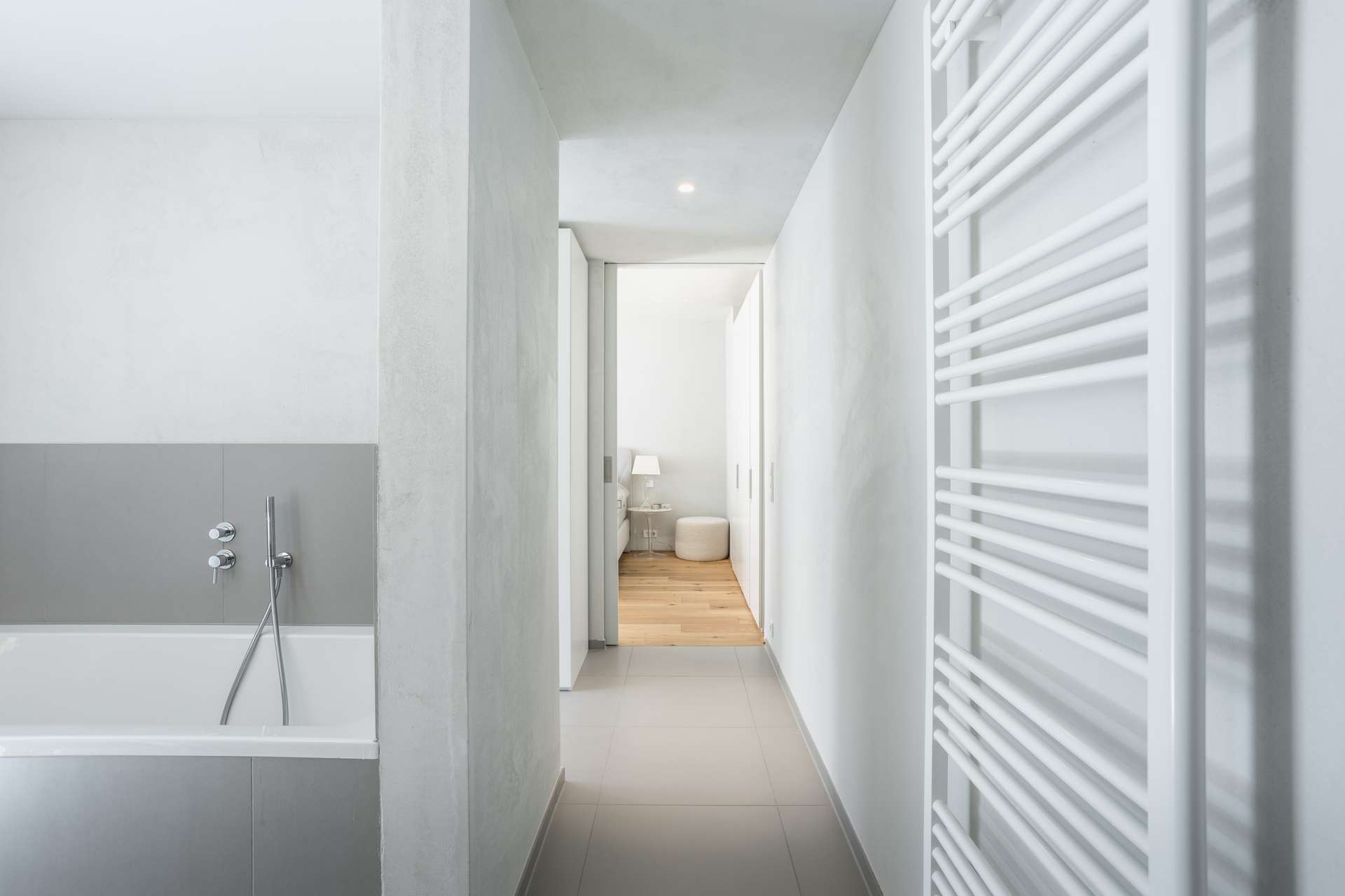 Bad Minimalistisches Badezimmer mit einer weißen Badewanne, einer Handbrause, grauen Fliesen und einem weißen Handtuchheizkörper. Der Flur führt zu einem Raum mit hellem Holzboden, einer kleinen Lampe und einem runden Hocker, der im Hintergrund zu sehen ist.
