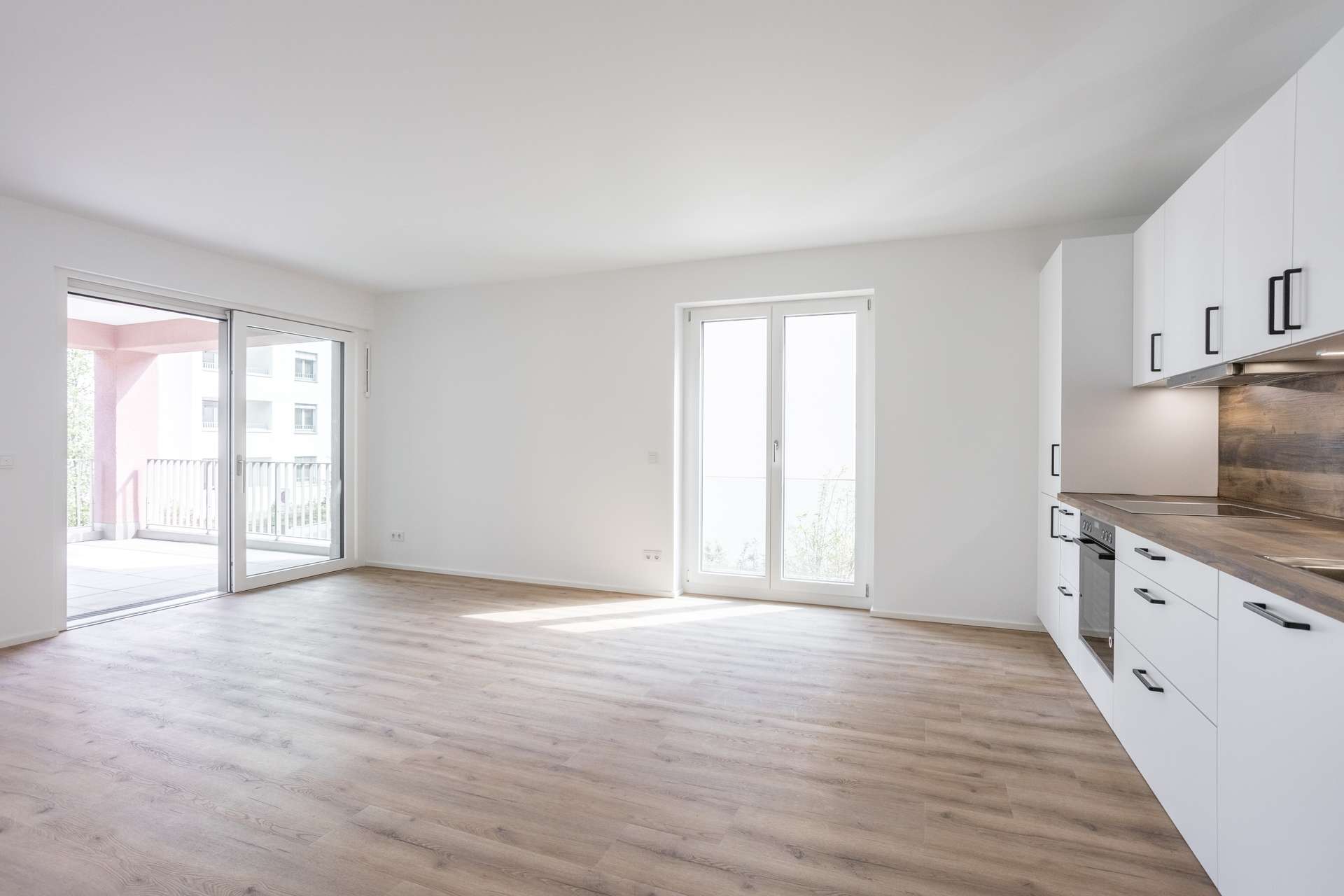 Wohn- und Essbereich Helle, leere, moderne Wohnung mit hellen Holzböden, weißen Wänden, großen Fenstern, einer Glasschiebetür zum Balkon und einer weißen Küche mit Holzakzenten auf der rechten Seite.