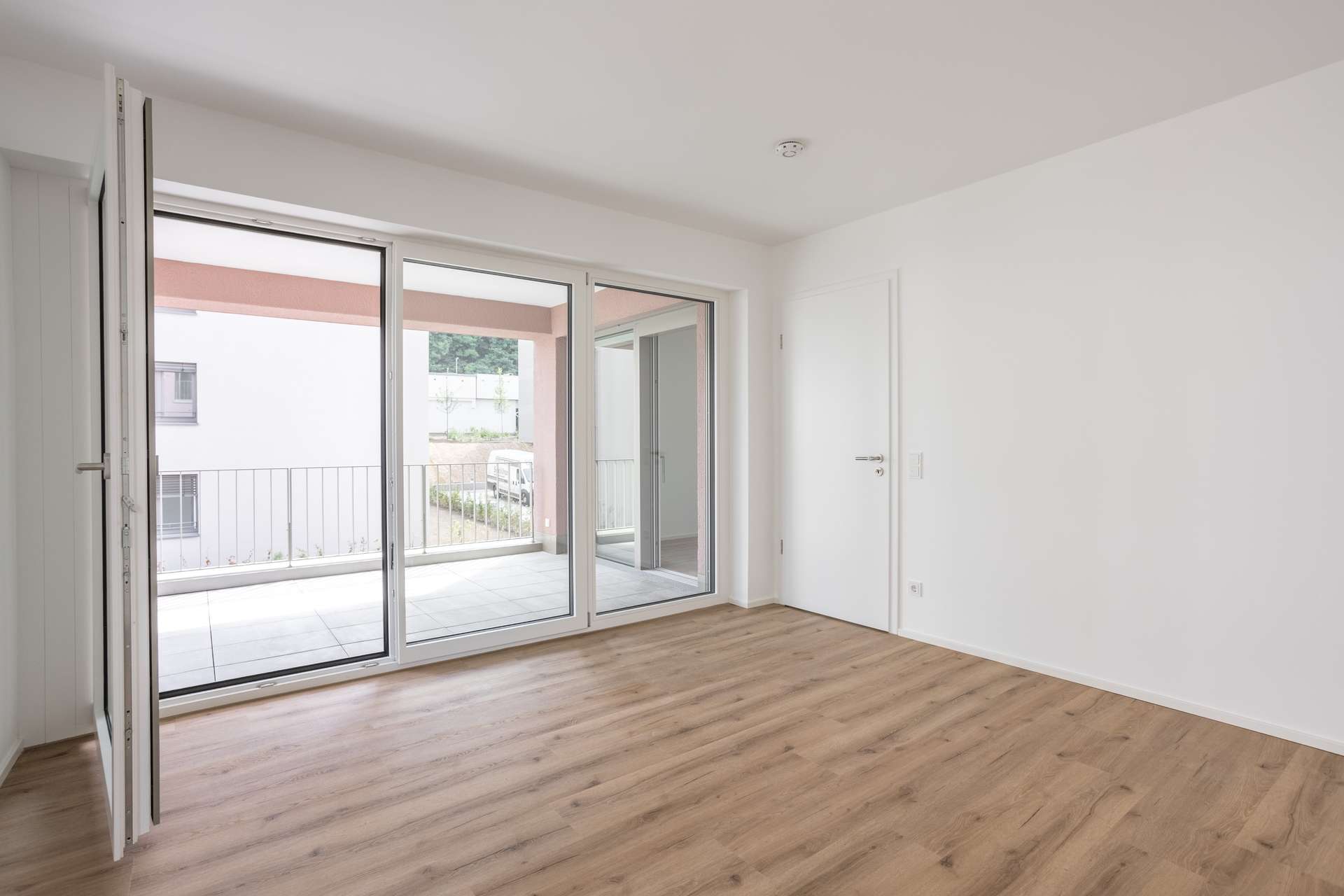 Schlafzimmer Ein heller, leerer Raum mit hellem Holzboden, weißen Wänden, einer Glastür und großen Fenstern, die auf einen Balkon führen. Der Balkon verfügt über ein Metallgeländer und bietet einen Blick auf andere Gebäude.