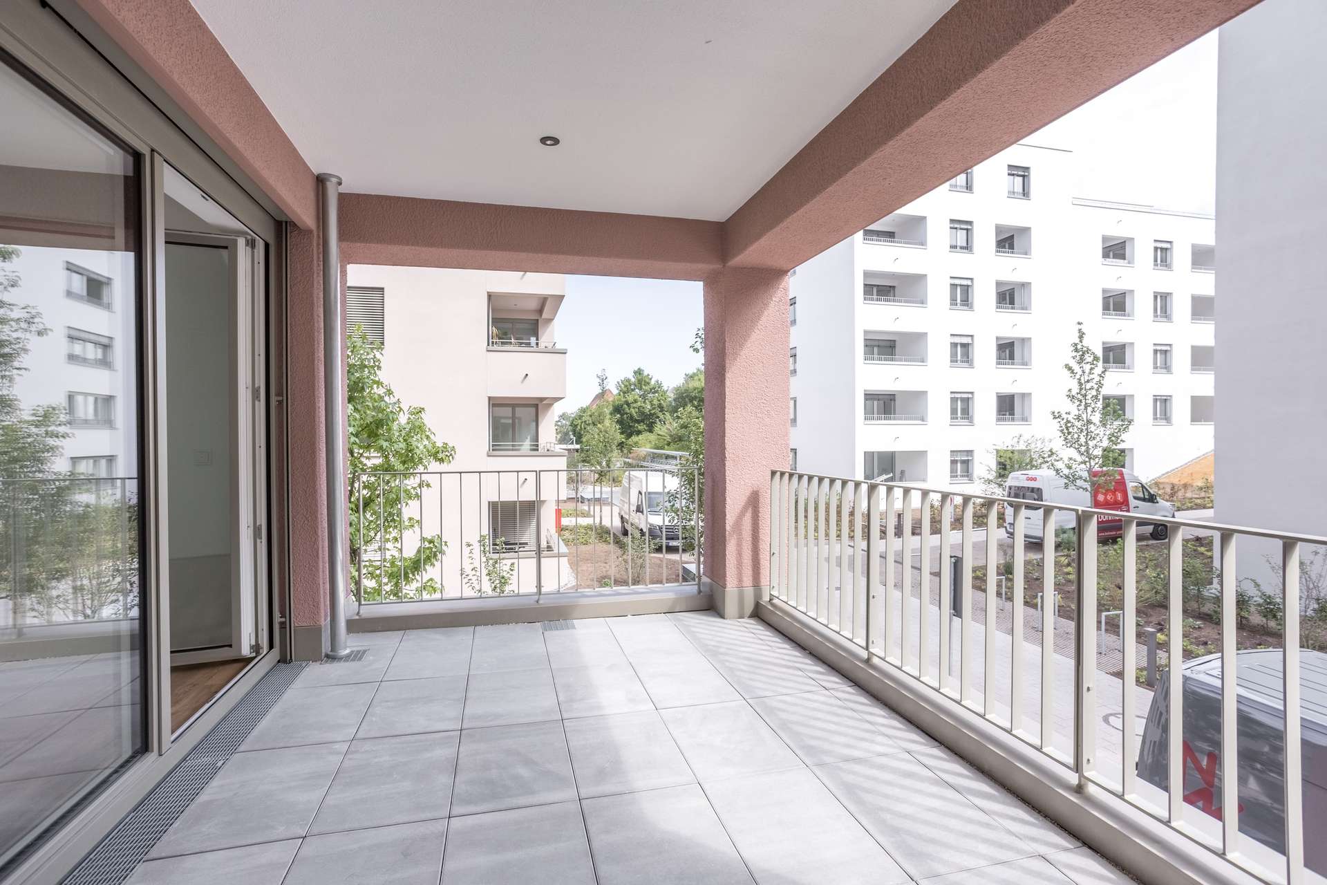 Loggia Geräumiger, gefliester Balkon mit weißem Geländer und Blick auf moderne weiße Wohnblocks, Bäume und ein geparktes Auto; Glasschiebetüren führen ins Innere.