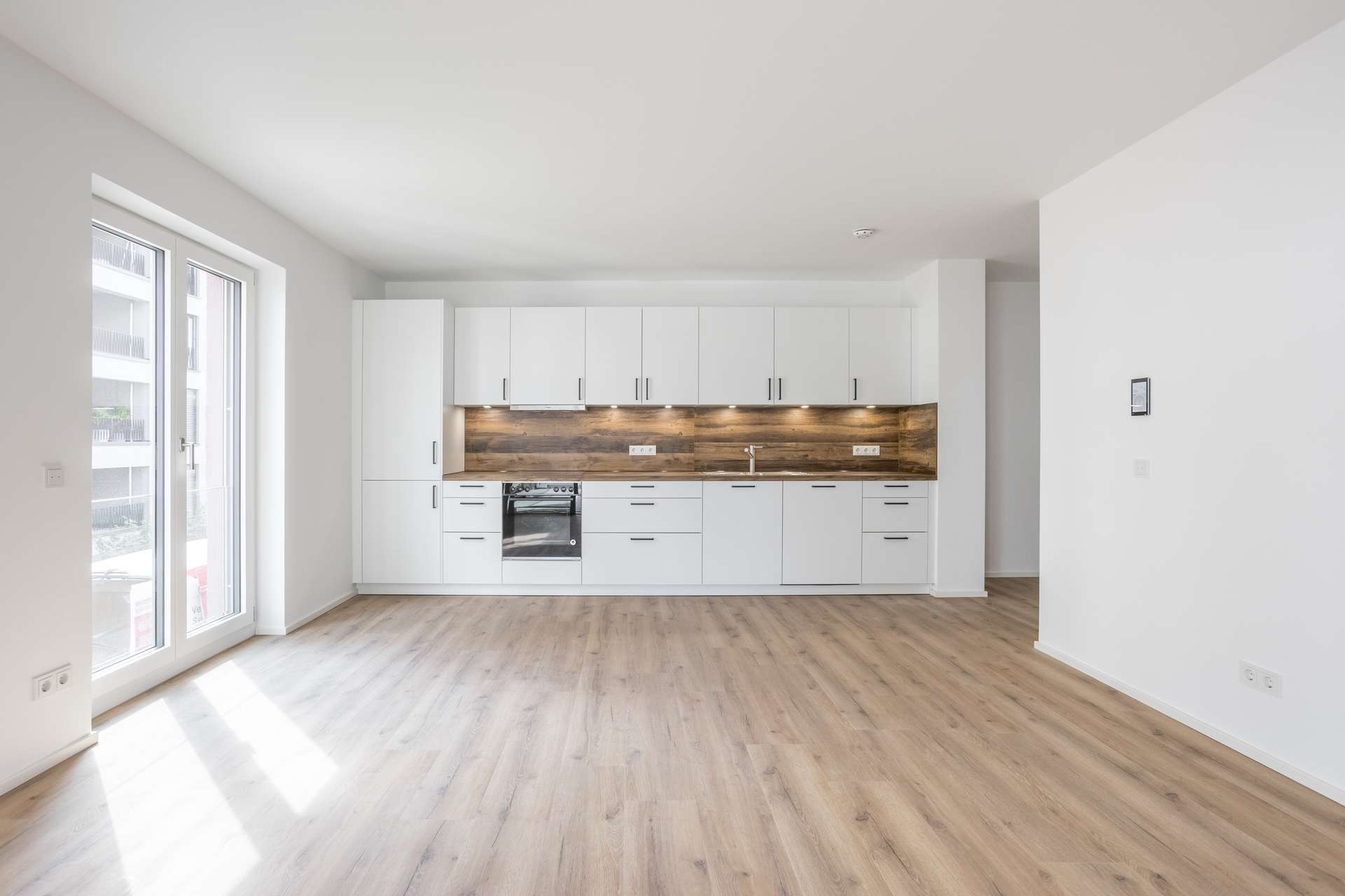 Küche Moderne, leere Küche mit weißen Schränken, einer Küchenrückwand aus Holz, einem Einbaubackofen und hellem Holzfußboden. Große Fenster auf der linken Seite lassen Tageslicht herein. Die Wände sind weiß und schaffen so einen hellen, aufgeräumten Raum.