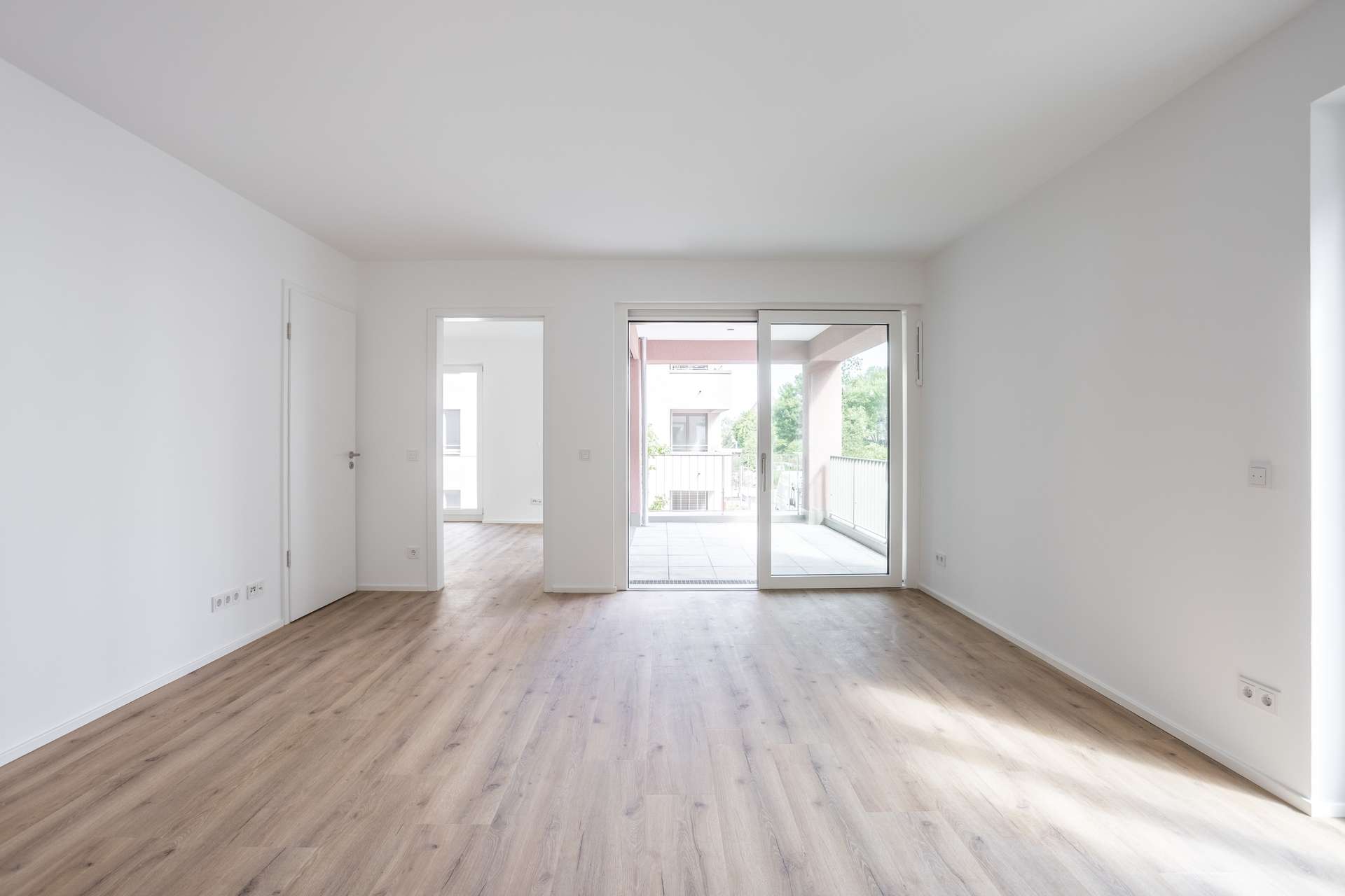 Wohn- und Essbereich Ein heller, leerer Raum mit hellem Holzboden, weißen Wänden und großen Glasschiebetüren, die auf einen Balkon führen. Zwei offene Durchgänge verbinden den Raum mit den angrenzenden Räumen. Natürliches Licht durchflutet den modernen, minimalistischen Raum.