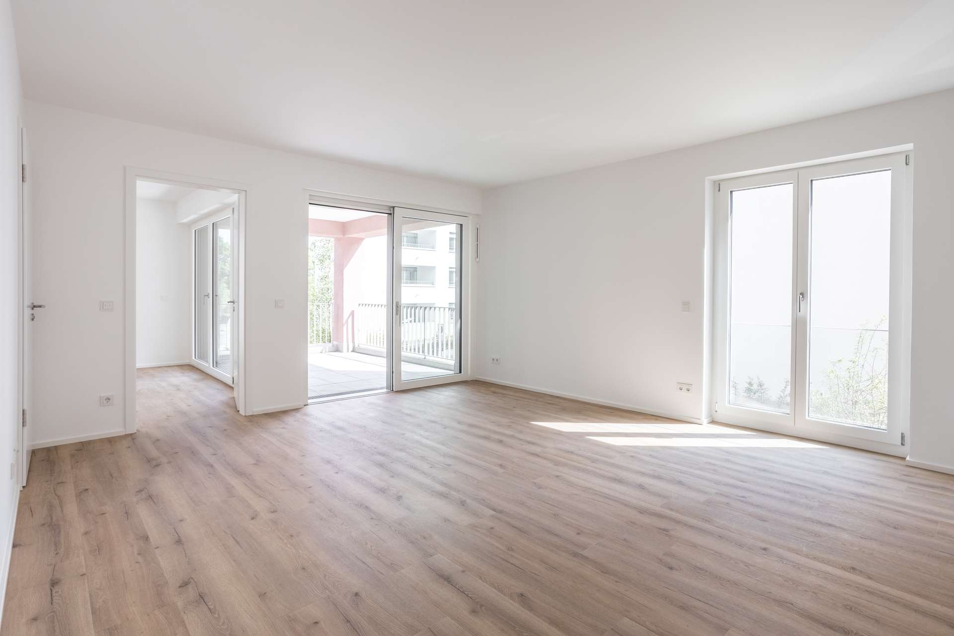 Wohn- und Essbereich Ein heller, leerer Raum mit hellem Holzboden, weißen Wänden, großen Fenstern und Glasschiebetüren, die auf einen Balkon führen. Natürliches Licht durchflutet den Raum und verleiht ihm eine luftige und großzügige Atmosphäre.